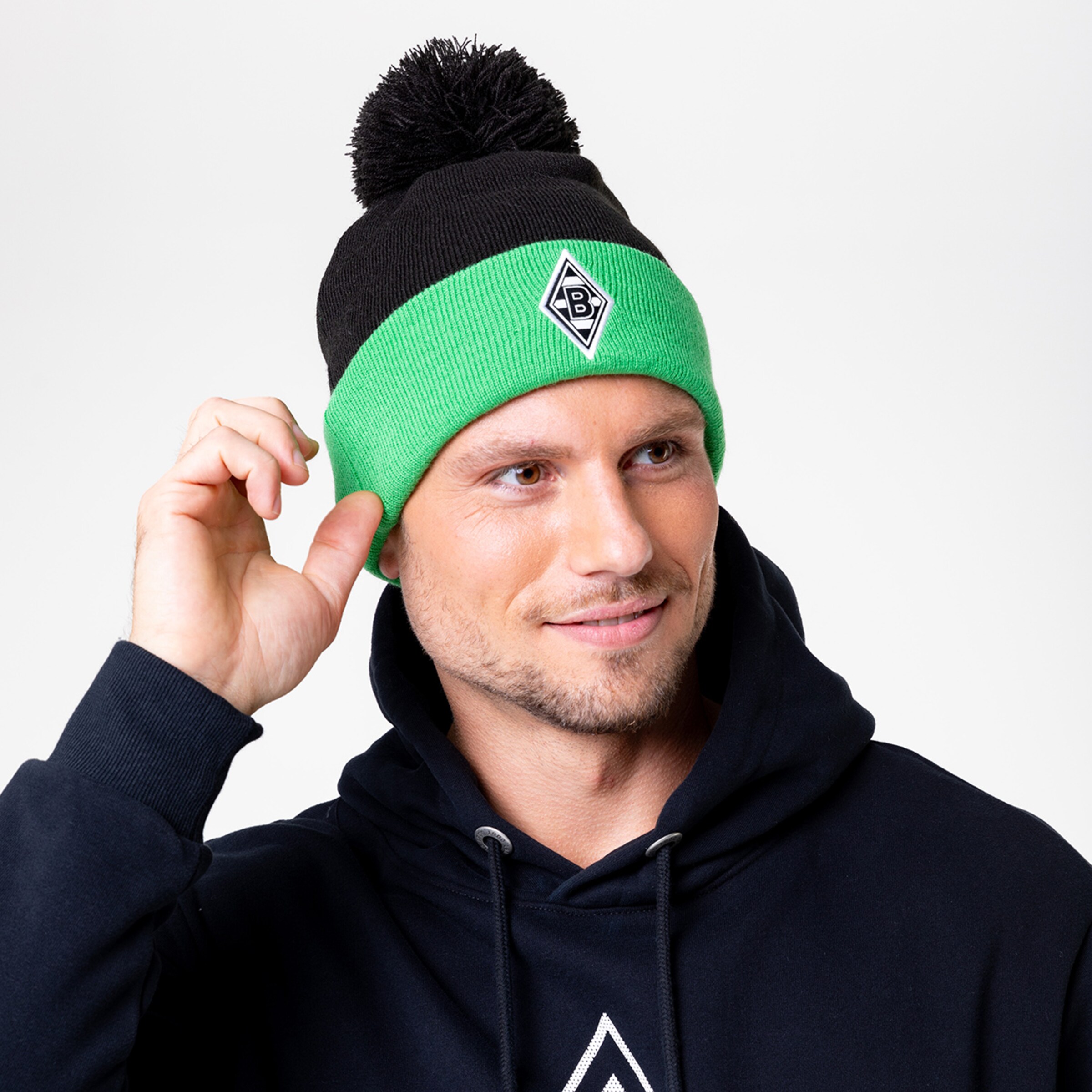 Mann mit Mütze in Schwarz und Grün trägt einen dunkelblauen Hoodie und lächelt.