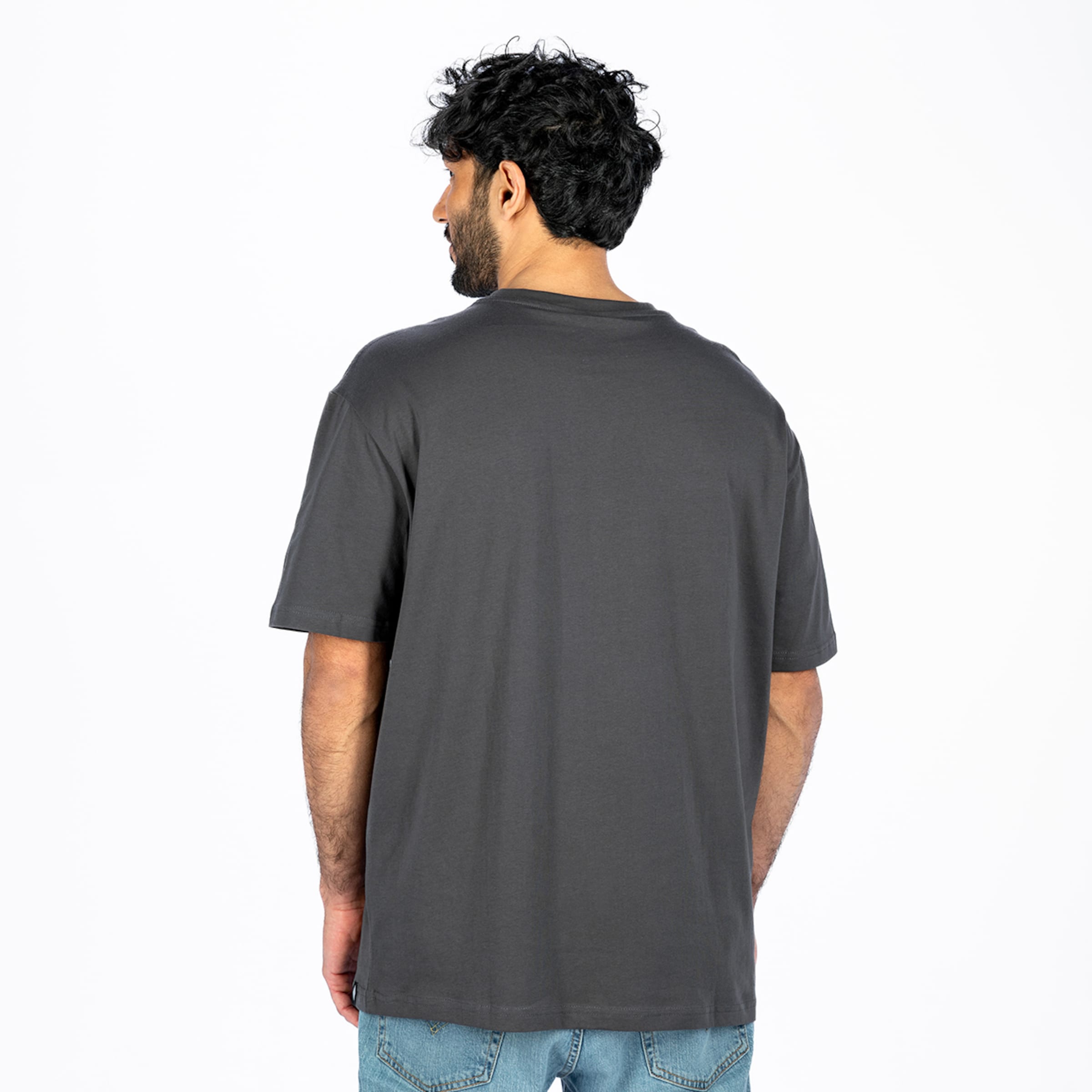 Rückansicht eines Mannes mit dunklem, lockigem Haar und Bart, der ein dunkelgraues, schlichtes T-Shirt und hellblaue Jeans vor einem weißen Hintergrund trägt. Sein Kopf ist nach links gedreht.