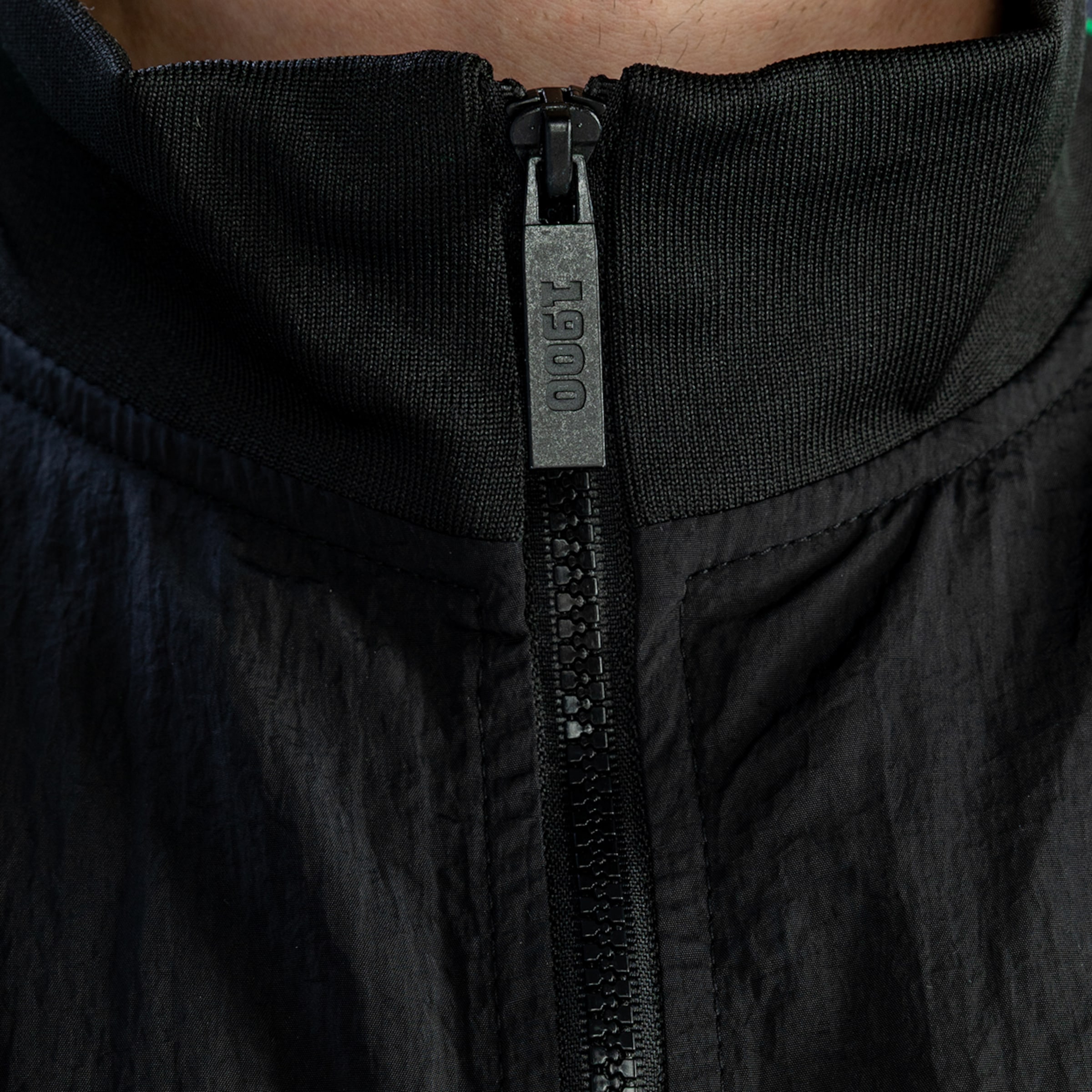 Nahaufnahme eines schwarzen Reißverschlusses. Der silberne Schieber zeigt die vertikale Prägung „1900“ vor einem gerippten schwarzen Kragen.