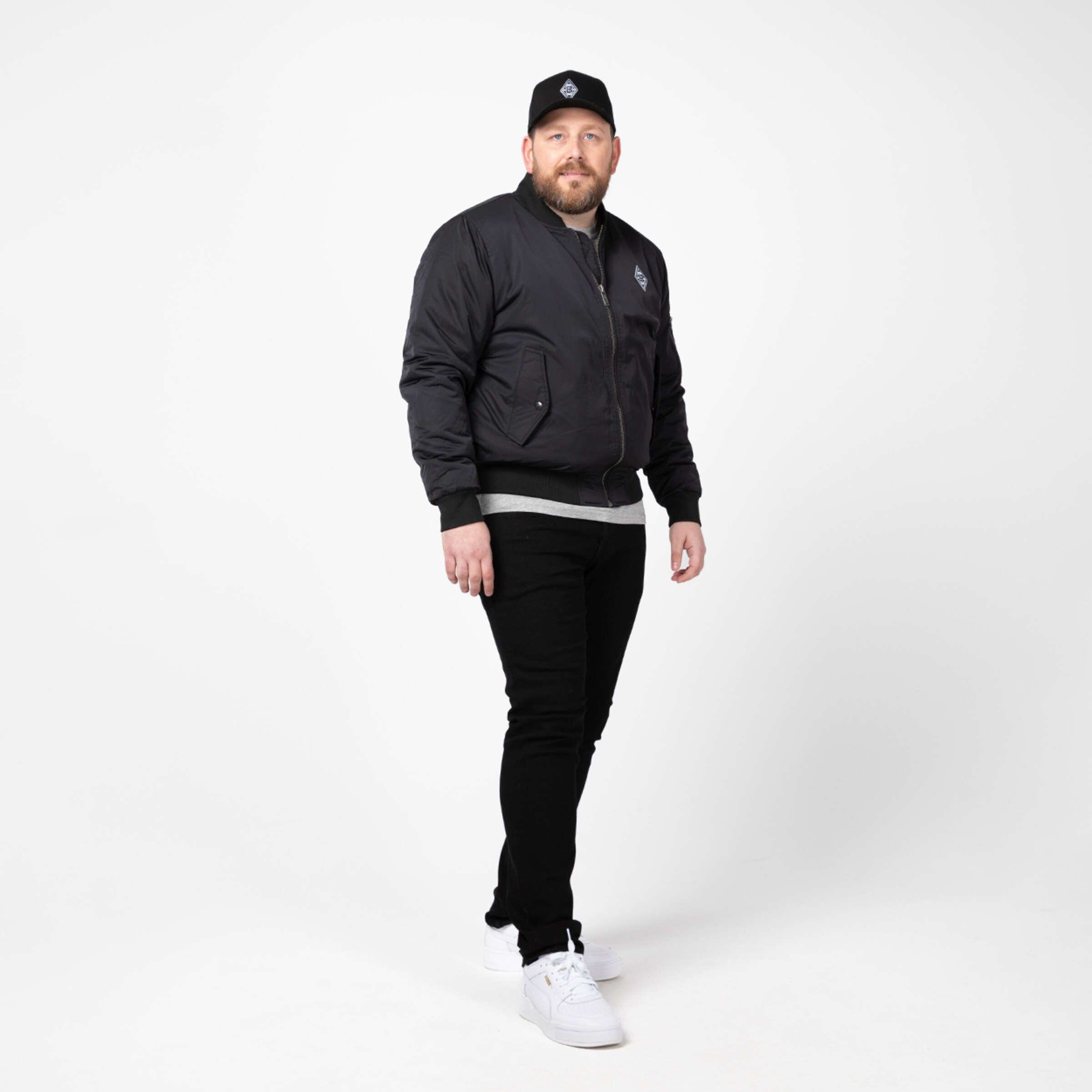 Man with a beard wearing a black jacket black pants and white sneakers.
