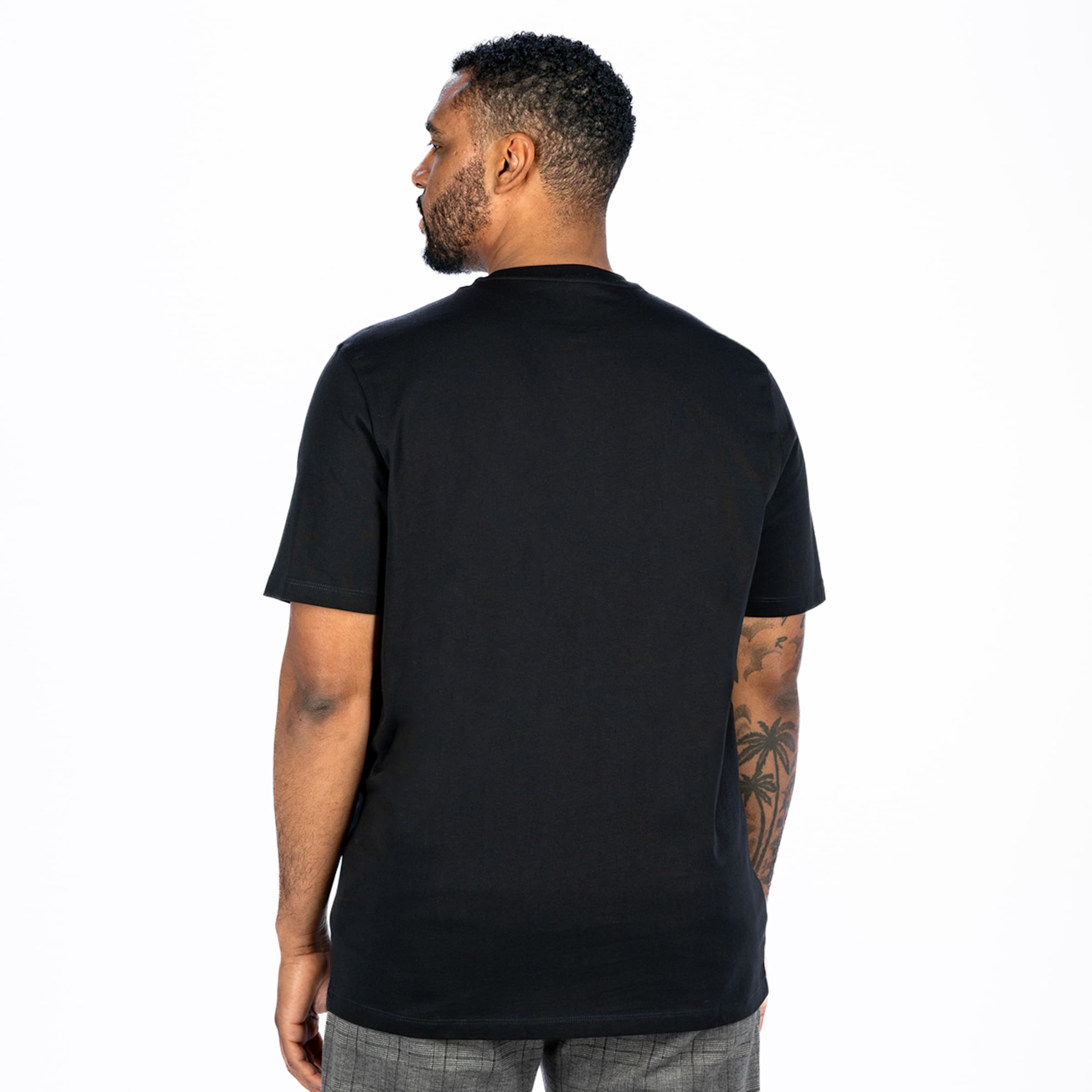 Rear view of a man wearing a black t-shirt and grey trousers against a white background. He has a tattoo on his right arm.