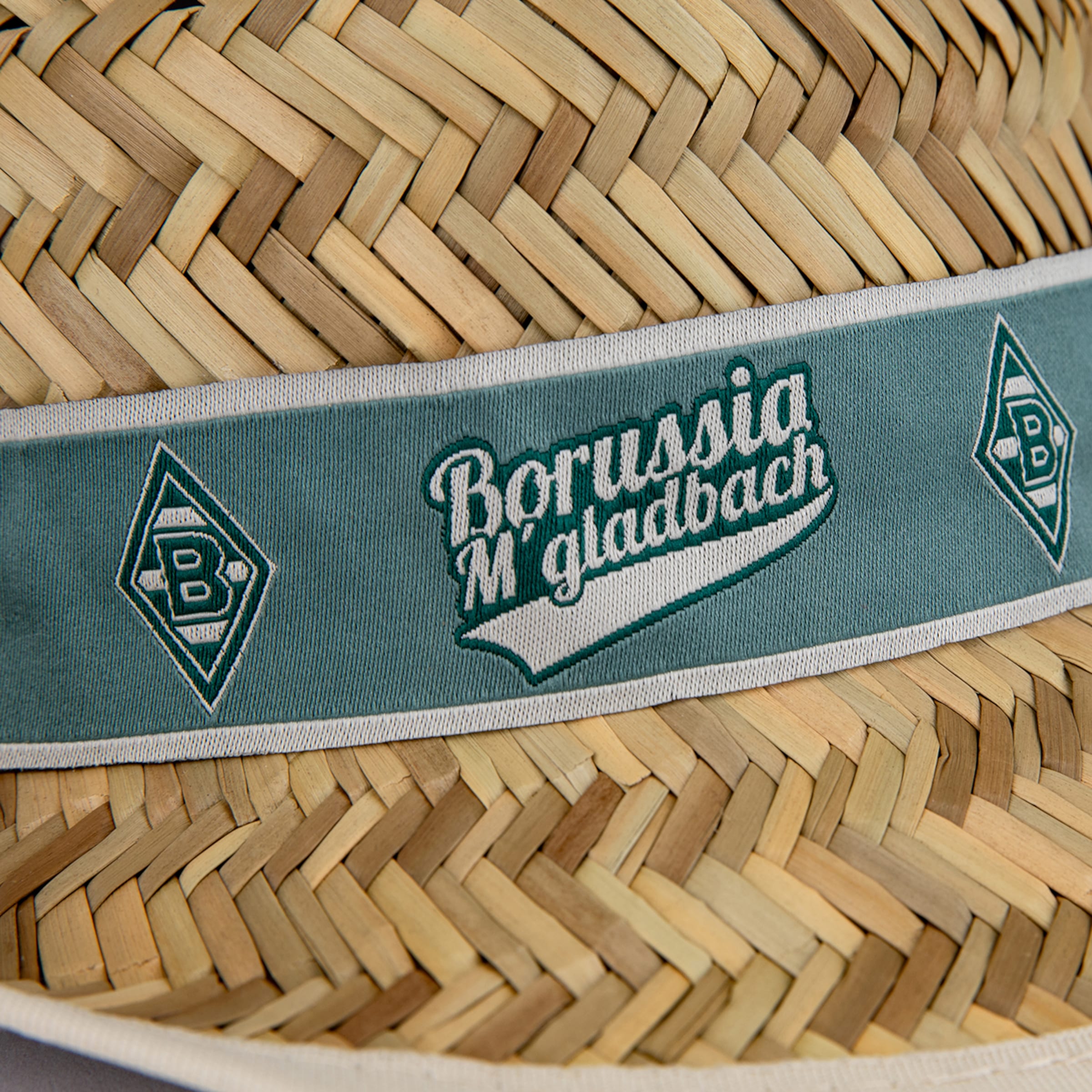 Nahaufnahme eines Strohhutes mit geflochtener Textur und einem grünen, bestickten Band. Das Band zeigt den Schriftzug „Borussia M'gladbach“ und rautenförmige Logos mit „B“s in Weiß und Grün.