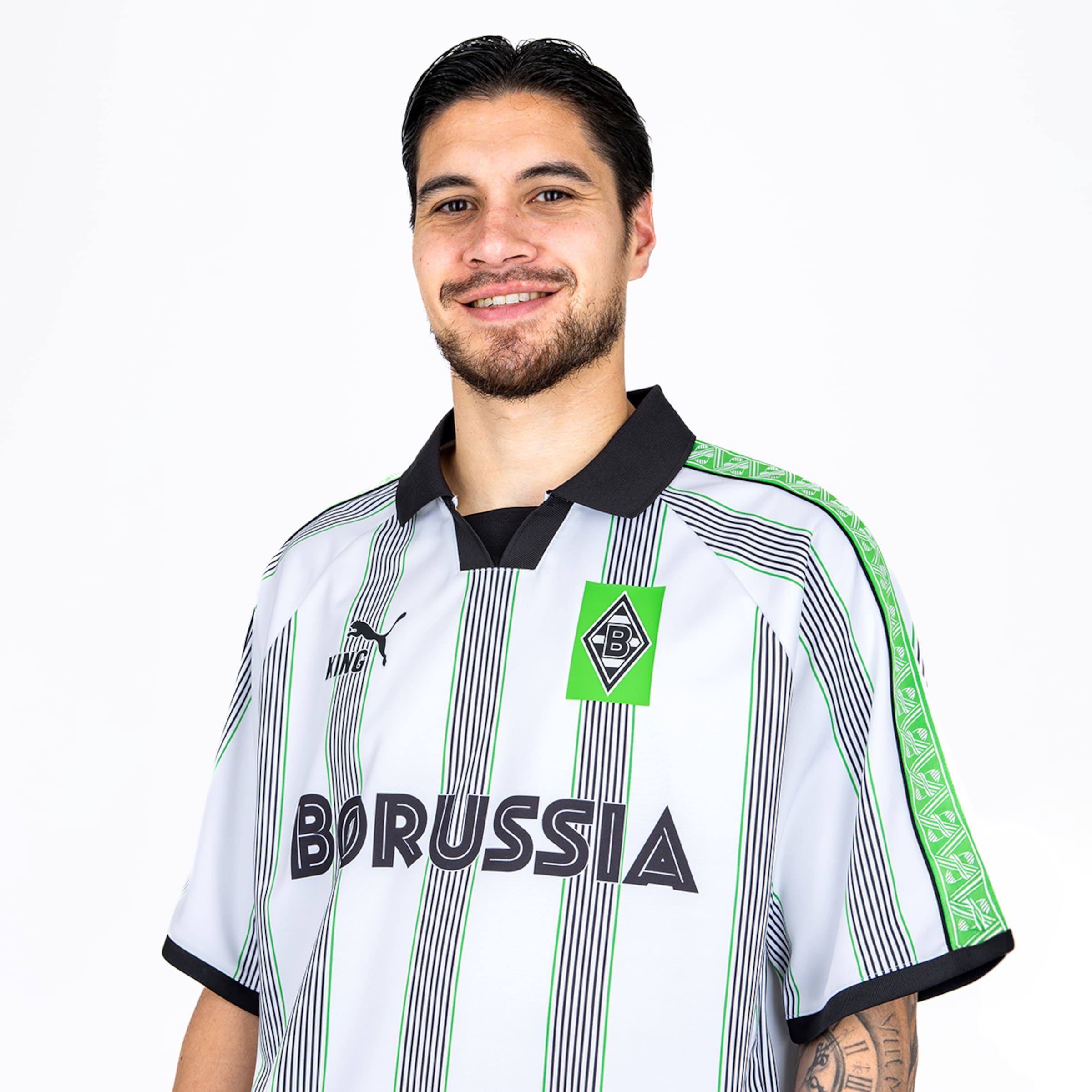 A smiling man wearing a white, green, and black striped Borussia Mönchengladbach football jersey against a plain white background.