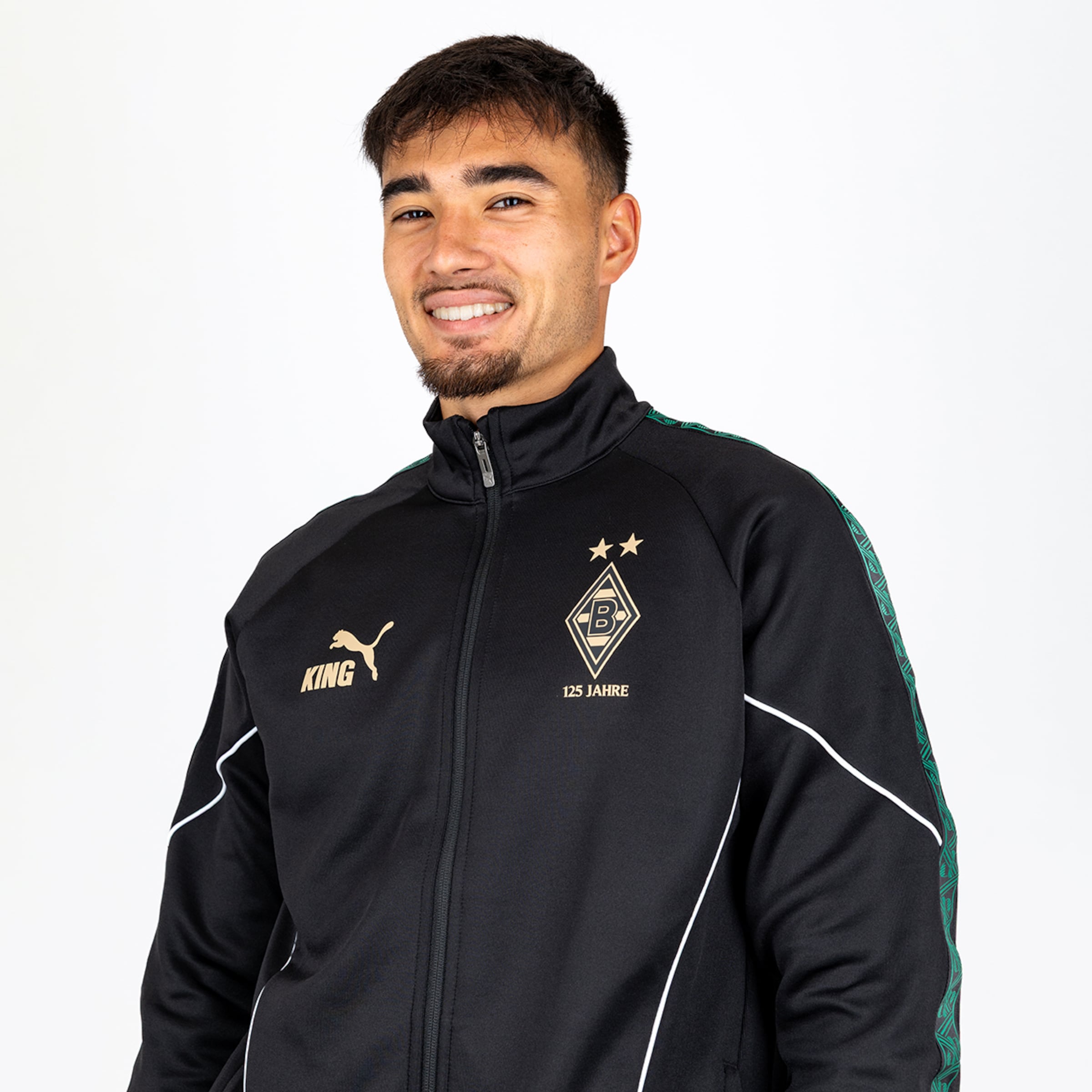 Lächelnder Mann in schwarzer Puma-Jacke mit Borussia Mönchengladbach Logo und „125 JAHRE“-Schriftzug vor weißem Hintergrund.