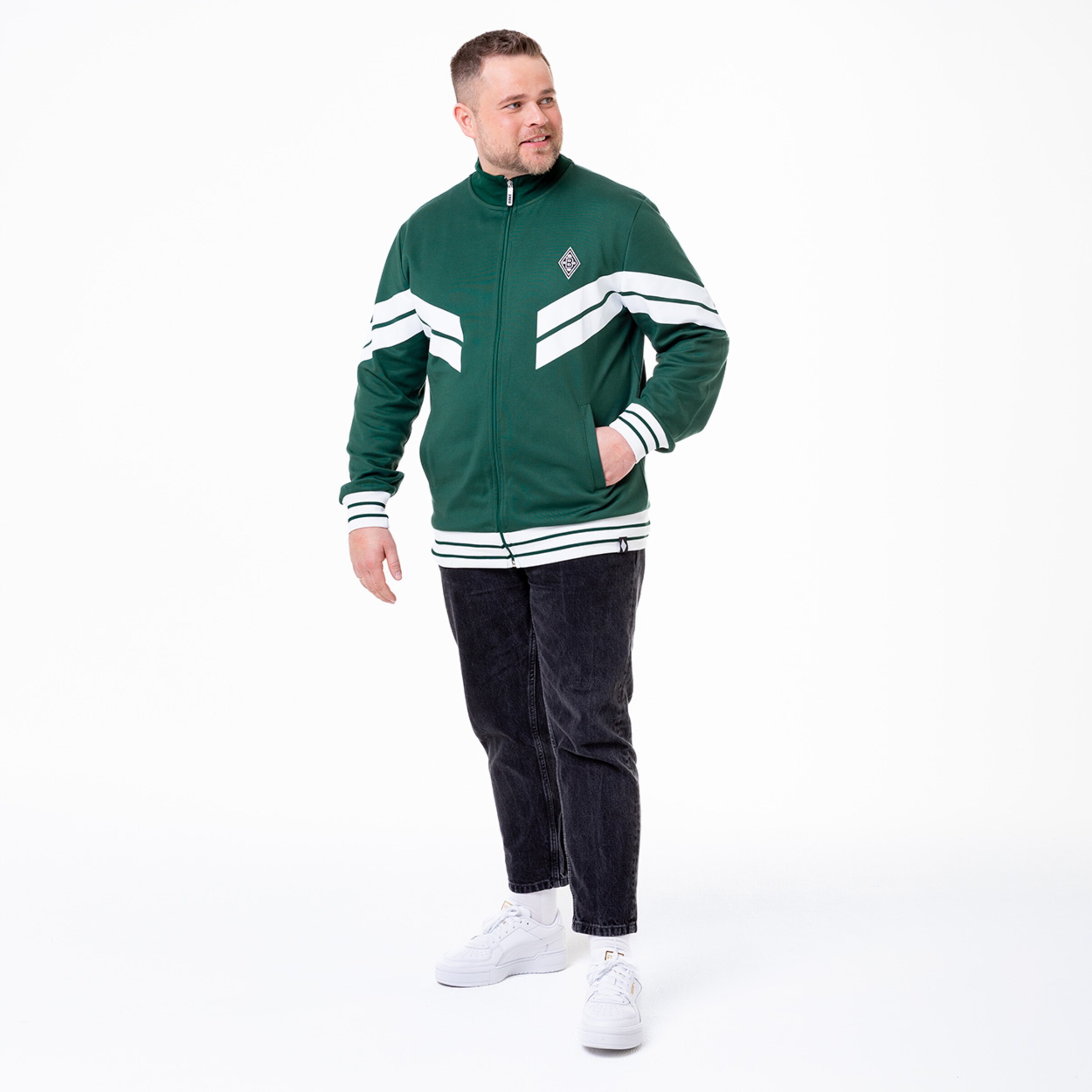 Mann in grünem Sportjackett mit weißen Streifen schwarze Jeans und weiße Sneakers.