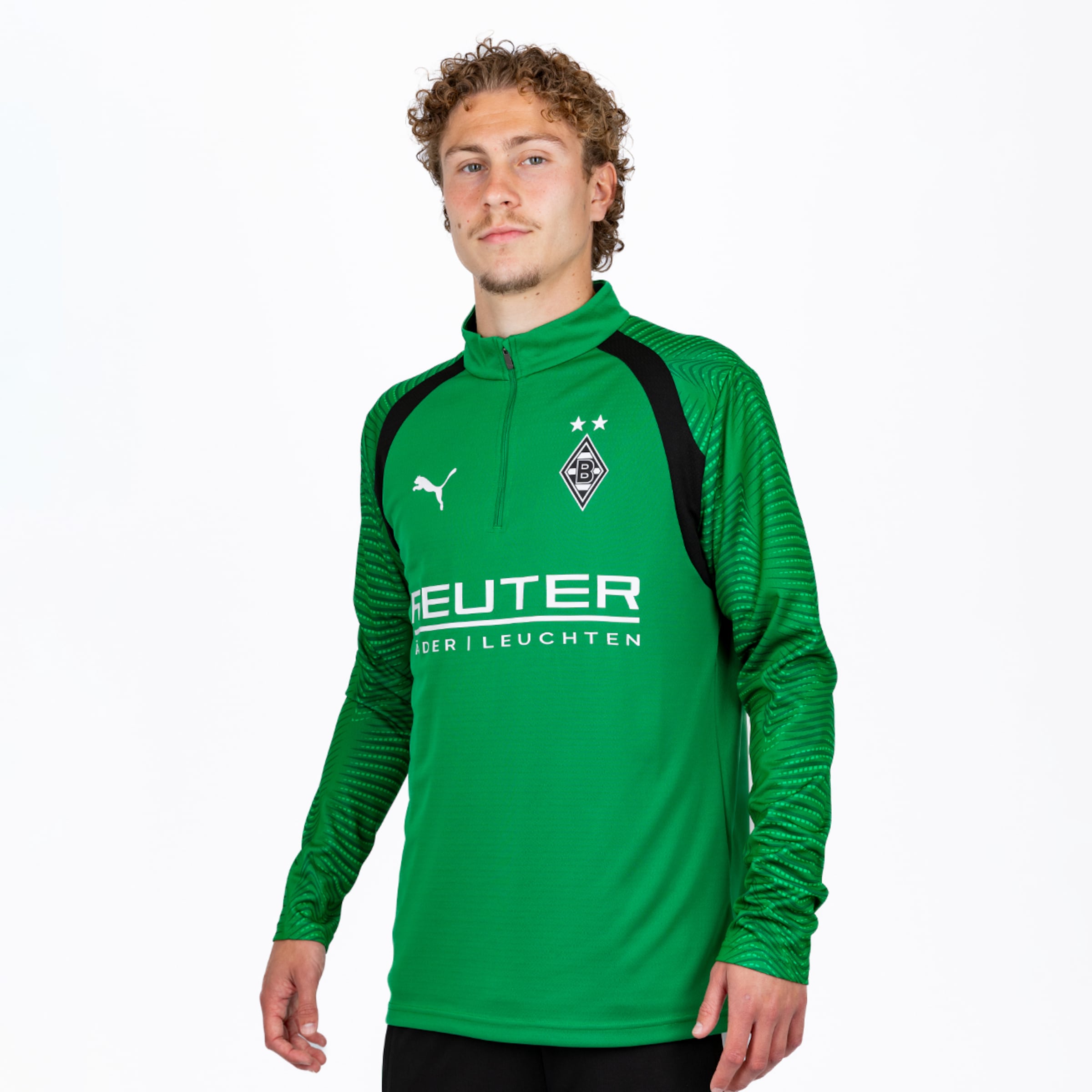 Ein männlicher Athlet mit lockigem Haar trägt ein grünes Borussia Mönchengladbach Puma-Trainingsoberteil mit Viertelreißverschluss, Vereinswappen mit zwei Sternen und dem Sponsor "REUTER | DER LEUCHTEN" vor weißem Hintergrund.