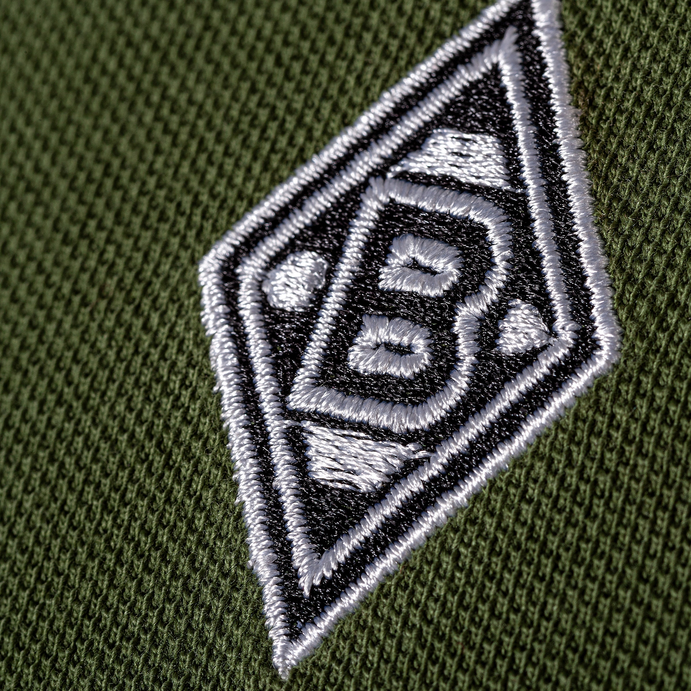Detailansicht des gestickten Borussia Mönchengladbach Logos (schwarz-weiße Raute mit „B“) auf grünem Strickstoff.