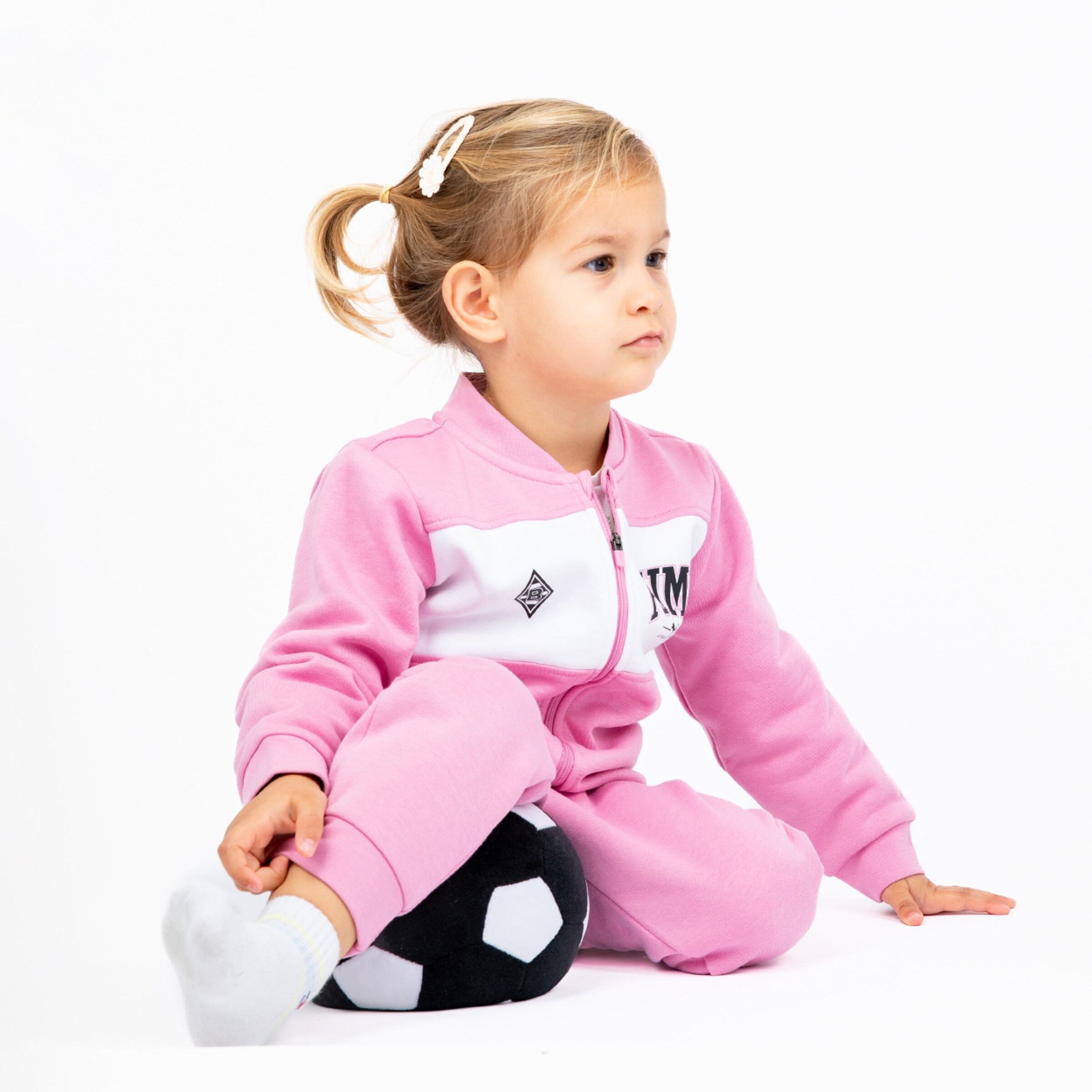 A young girl in a pink tracksuit sits on a soccer ball looking thoughtful.