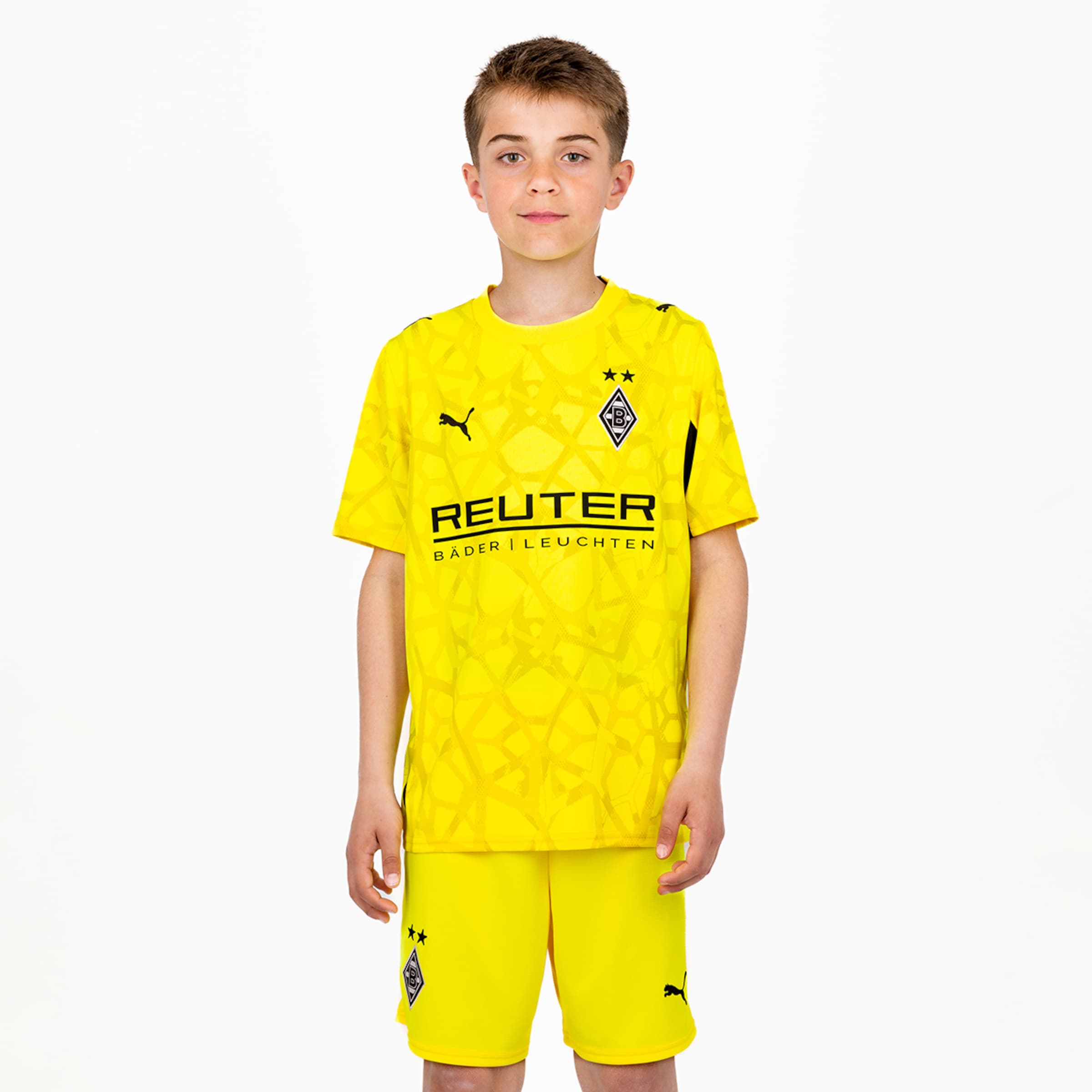 Ein Junge im gelben Borussia Mönchengladbach Trikot mit Reuter-Logo und passenden Shorts auf weißem Hintergrund.