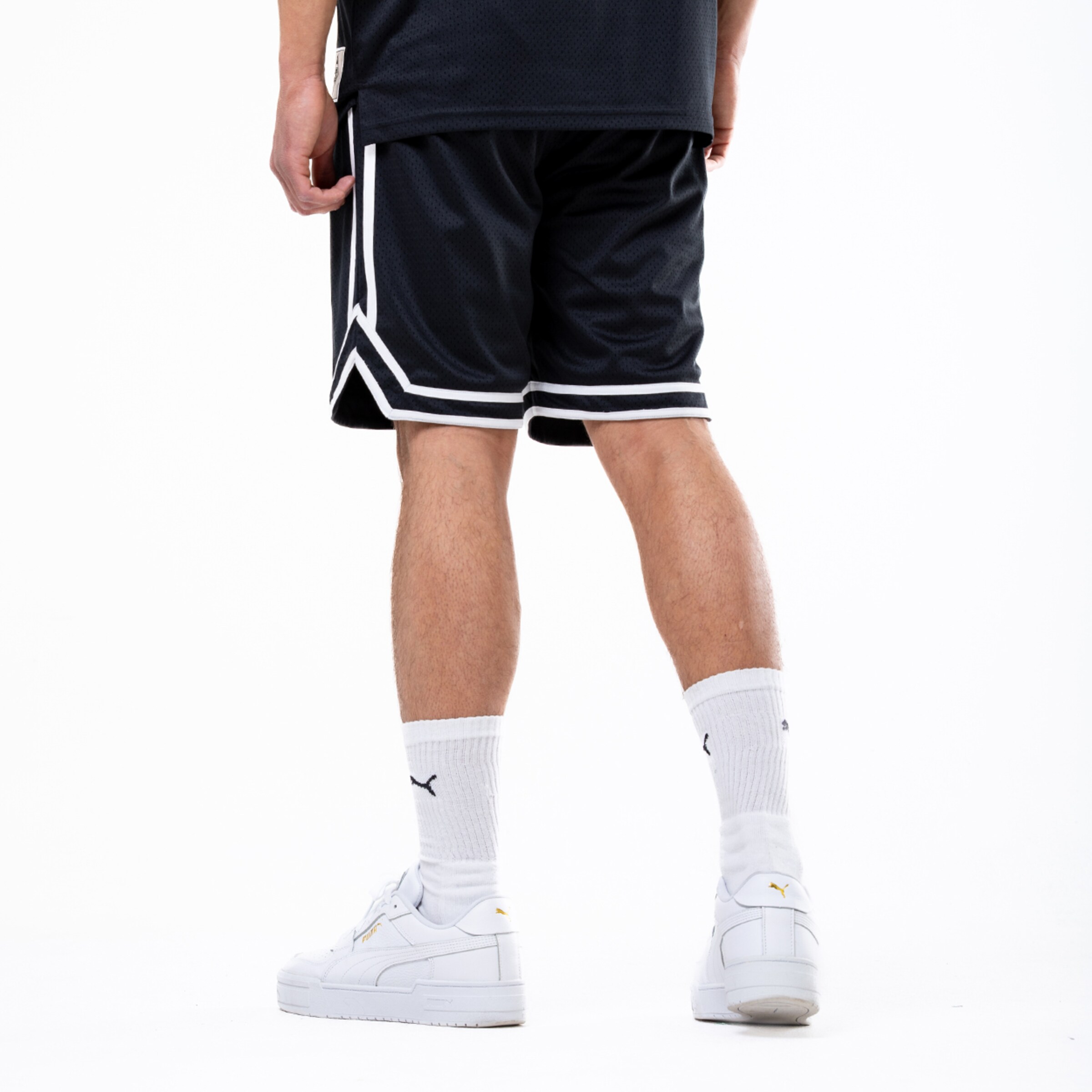 Rückansicht einer Person in schwarzen Basketball-Shorts mit weißen Streifen, weißen Sportsocken und weißen Sneakern vor weißem Hintergrund.