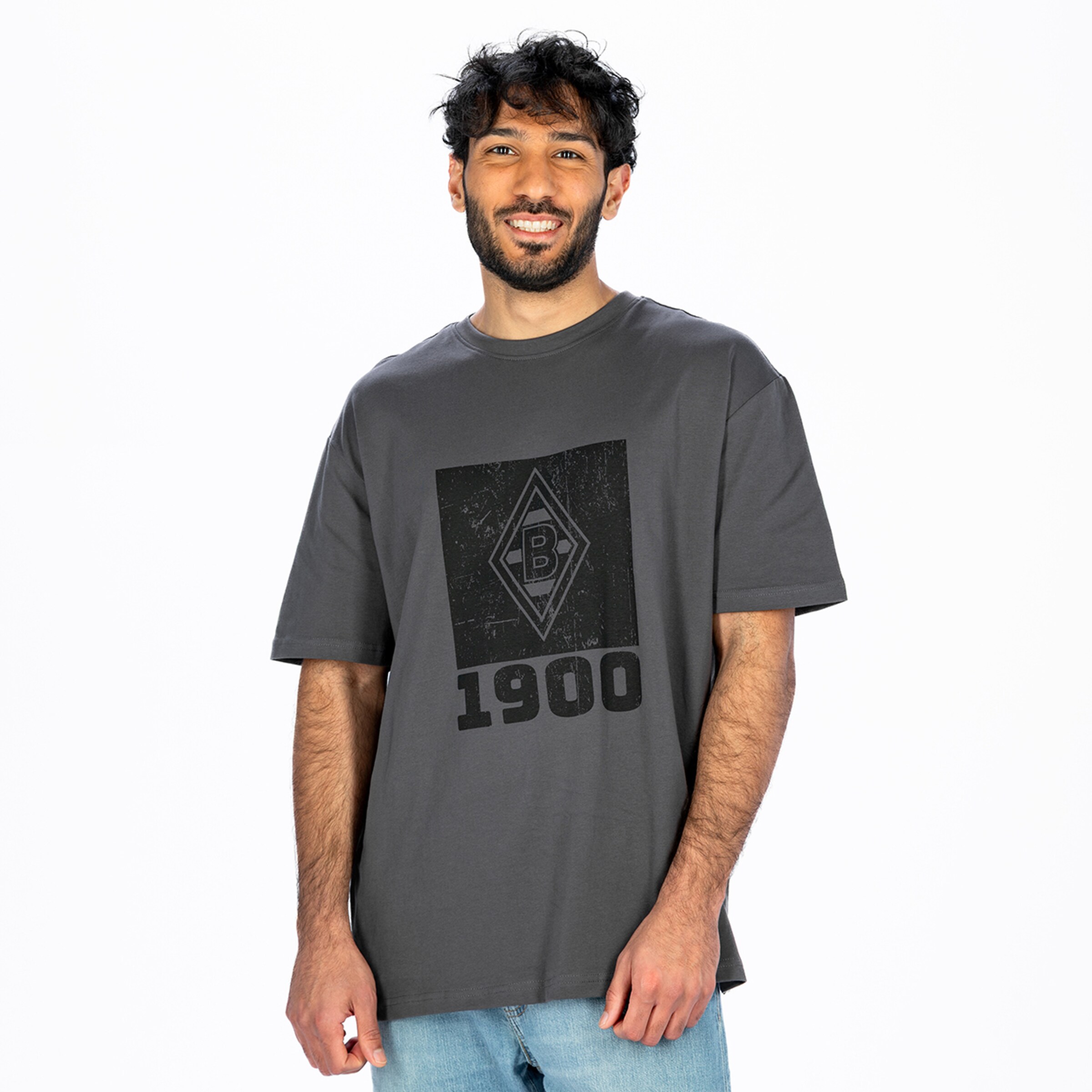 Ein lächelnder Mann mit dunklem Lockenhaar und Bart, trägt ein graues T-Shirt mit einem schwarzen, abgenutzten Borussia Mönchengladbach Logo und der Jahreszahl „1900“ sowie hellblaue Jeans vor weißem Hintergrund.