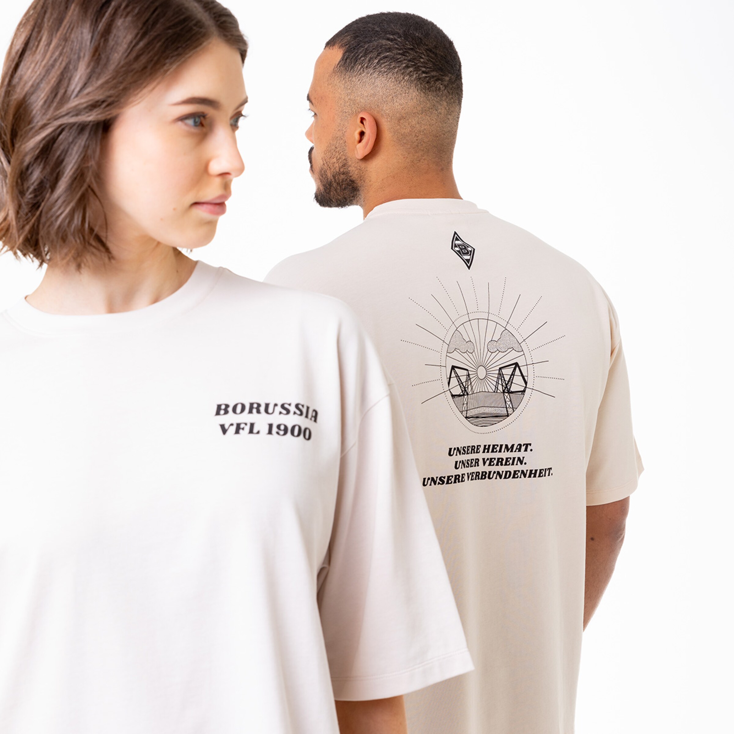 A woman and a man wearing T-shirts. The womans shirt features BORUSSIA VFL 1900 on the front. The mans shirt displays a graphic design on the back with the text UNSER HEIMAT. UNSER VERBINDENHEIT.