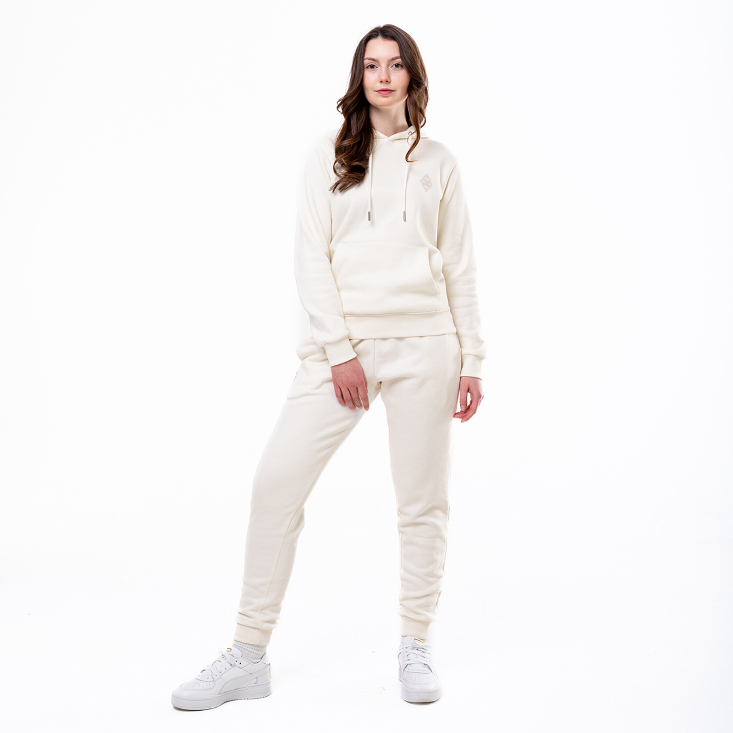 Eine Frau steht in einem hellen hellbeigen Jogginganzug mit Kapuze und weißen Sneakers.