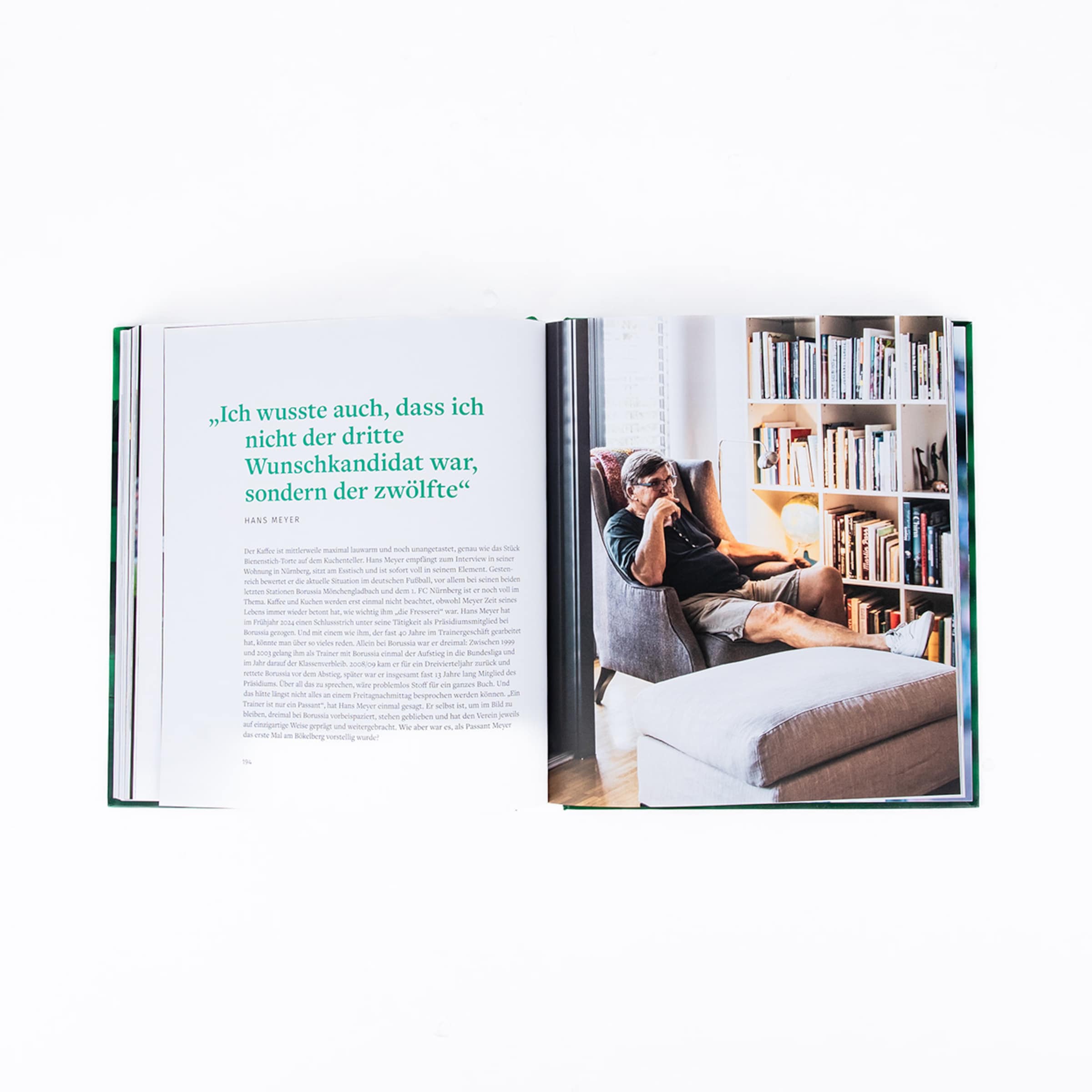 Ein Mann sitzt in einem Sessel und liest ein Buch umgeben von Regalen voller Bücher.