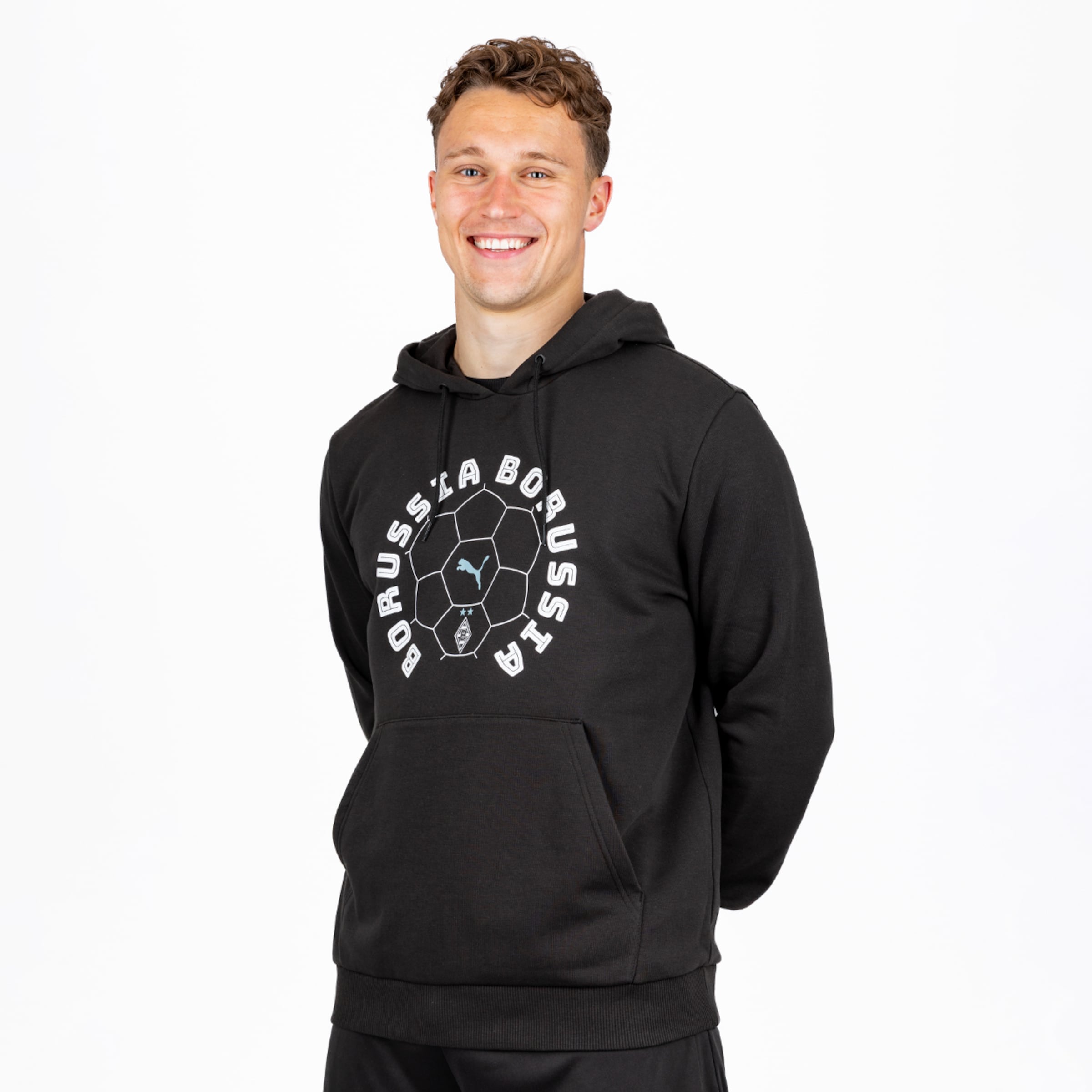 Lächelnder Mann mit braunen Locken in schwarzem Borussia Mönchengladbach Hoodie mit Fußball-Design, „BORUSSIA“-Schriftzug, Puma- und Vereinslogos vor weißem Hintergrund.