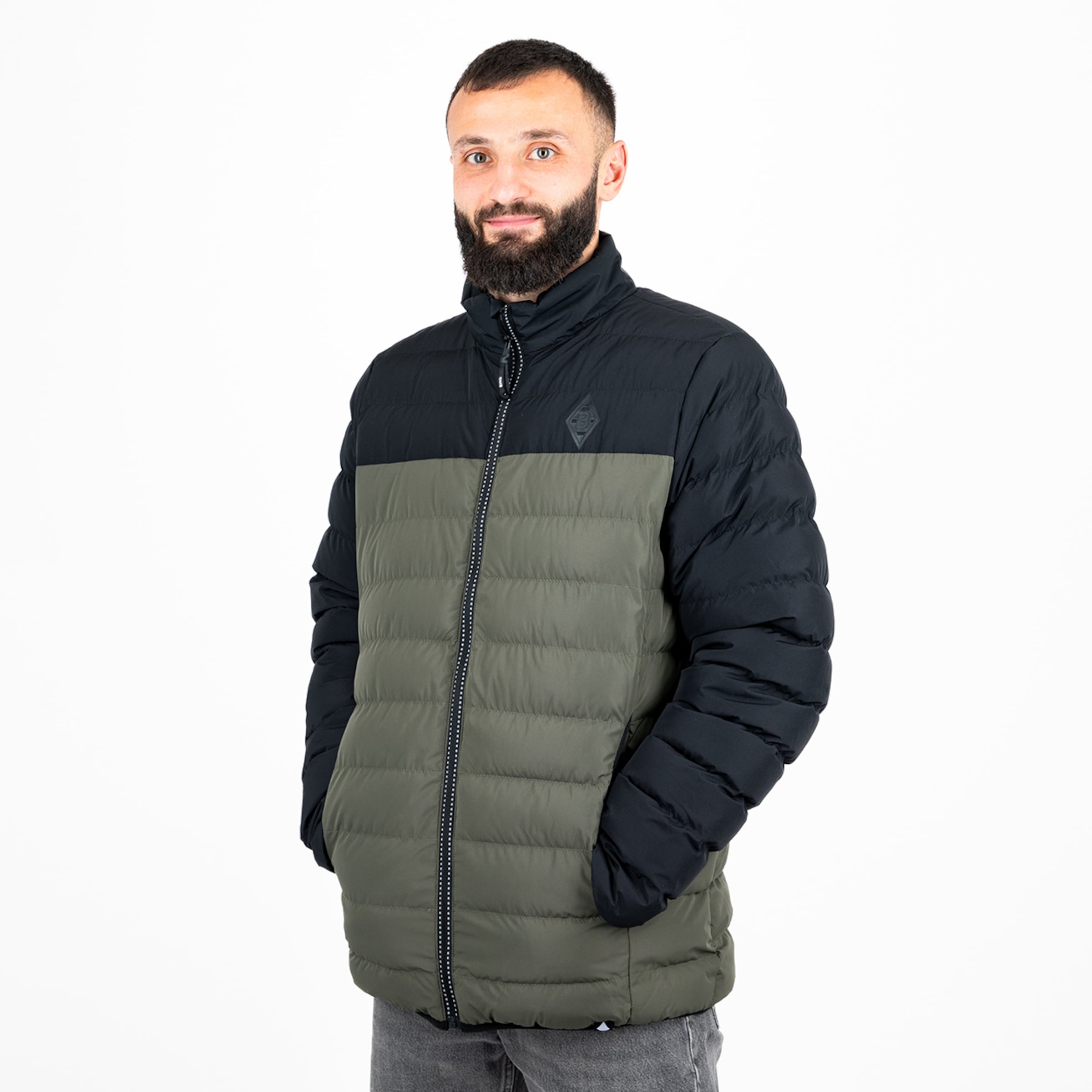 Ein Mann trägt eine dunkelgrüne und schwarze Jacke mit Reißverschluss und hat die Hände in den Taschen.