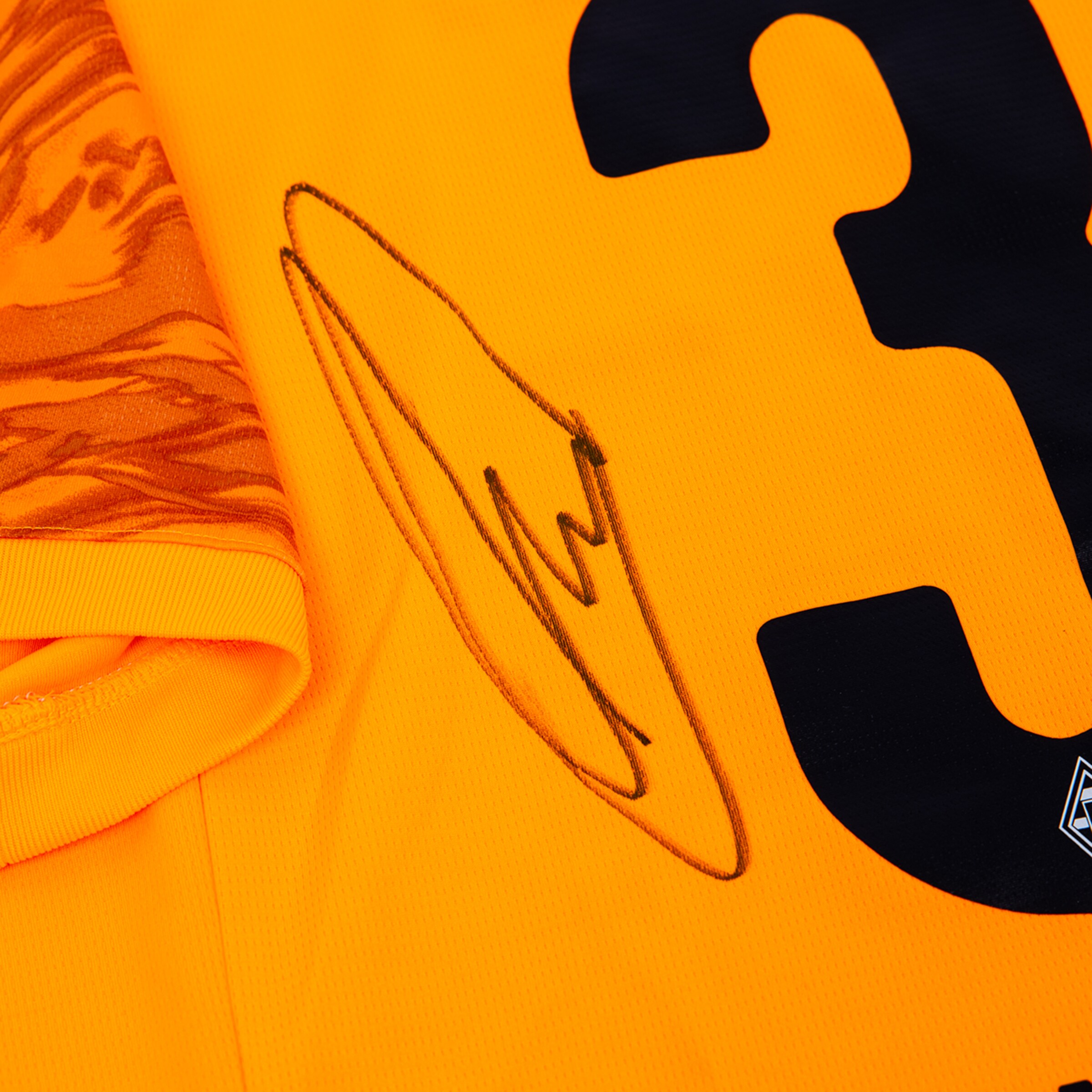 Close-up of an orange jersey with a black signature next to the number 3 and a patterned sleeve on the left.