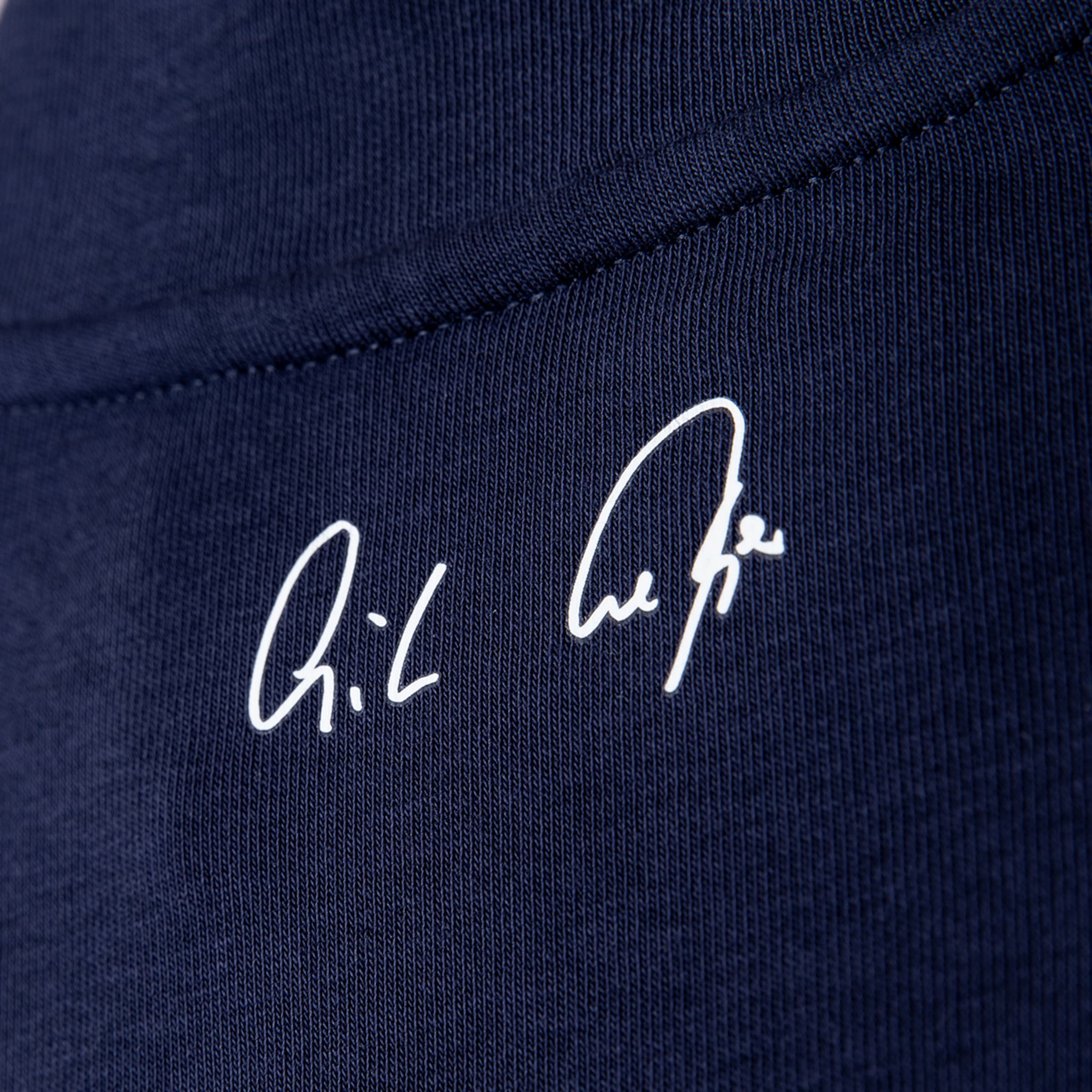 Weiße, kursive Signatur auf dunkelblauem Stoff mit sichtbarer Textur in einer Nahaufnahme.