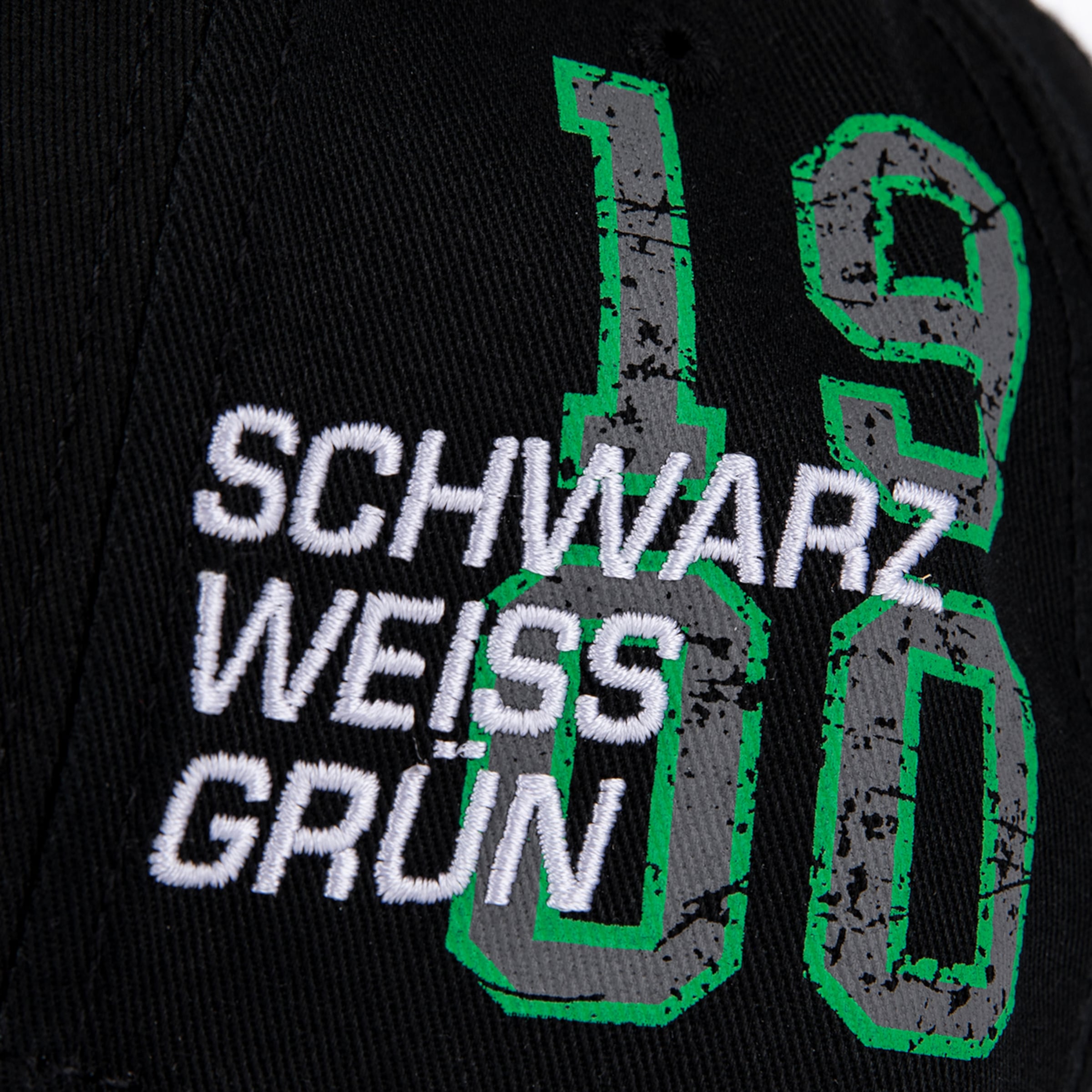 Weißer Stick „SCHWARZ WEISS GRUN“ auf schwarzem Stoff vor großen, grau-grün umrandeten Zahlen „1900“ im Distressed-Look.