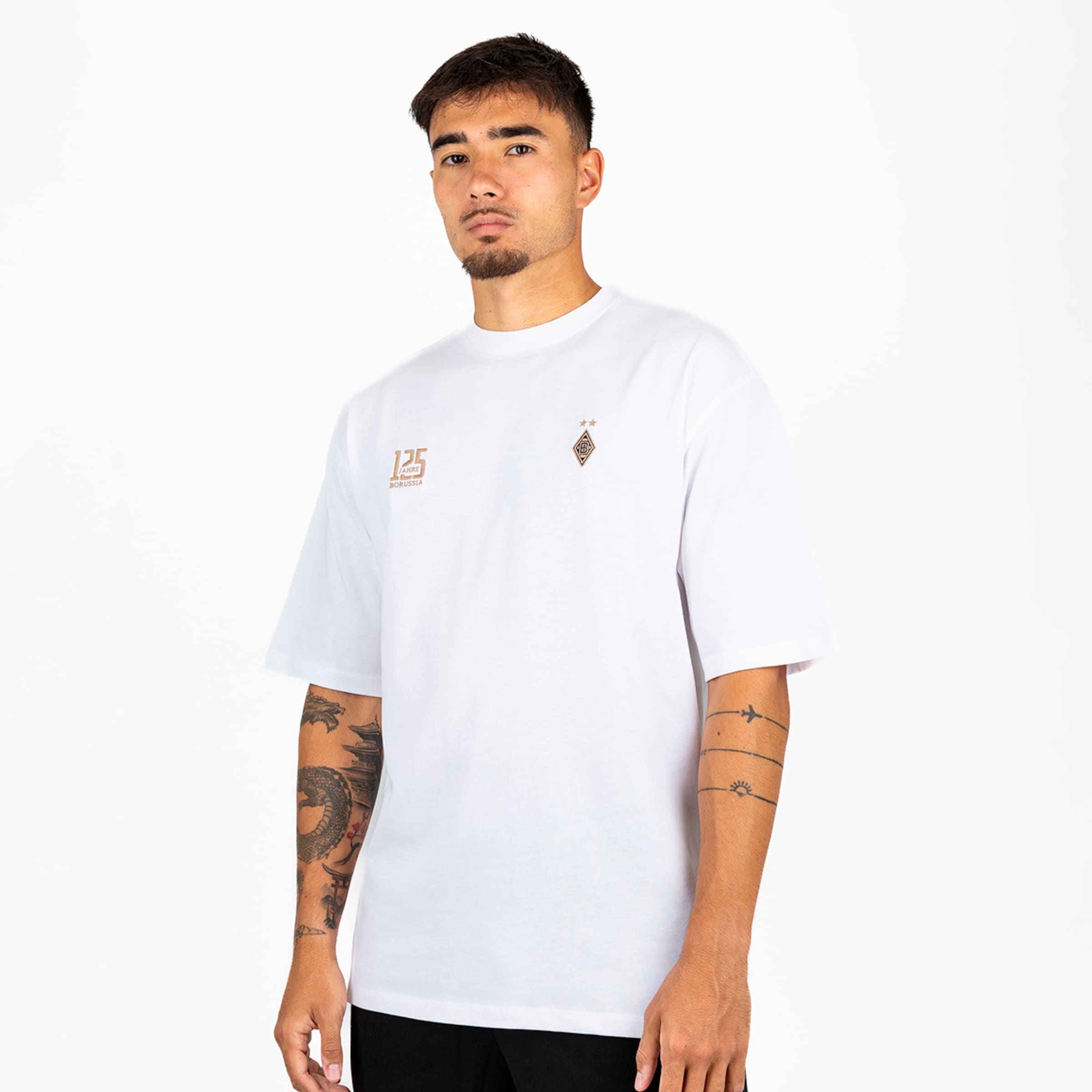 Mann in weißem Oversize-T-Shirt mit zwei kleinen goldenen Logos auf der Brust vor weißem Hintergrund. Er hat Tattoos an beiden Armen.
