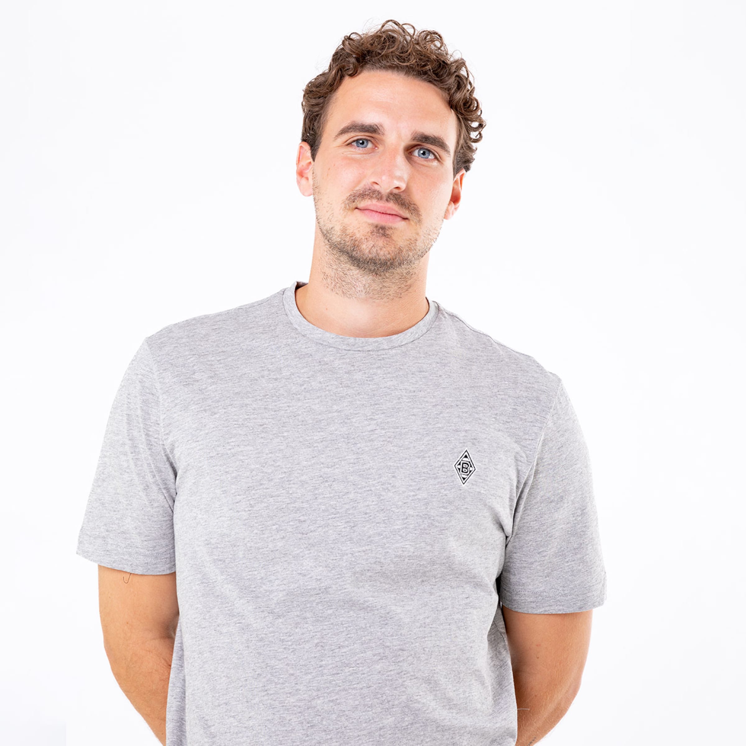 Junger Mann mit lockigem braunem Haar trägt ein graues T-Shirt mit einem kleinen Aufdruck.