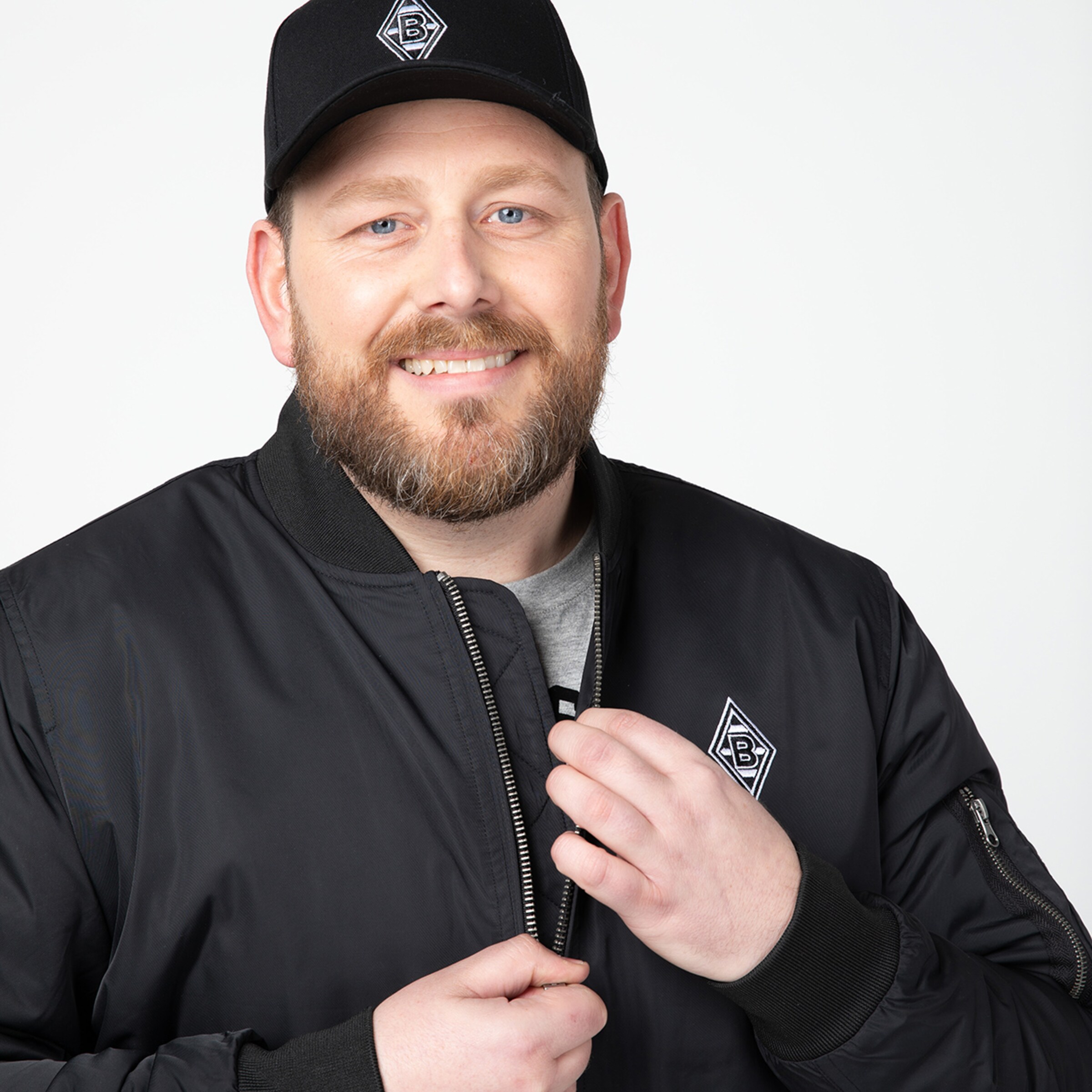 Man with a beard smiling holding the zipper of his jacket wearing a black baseball cap with a logo.