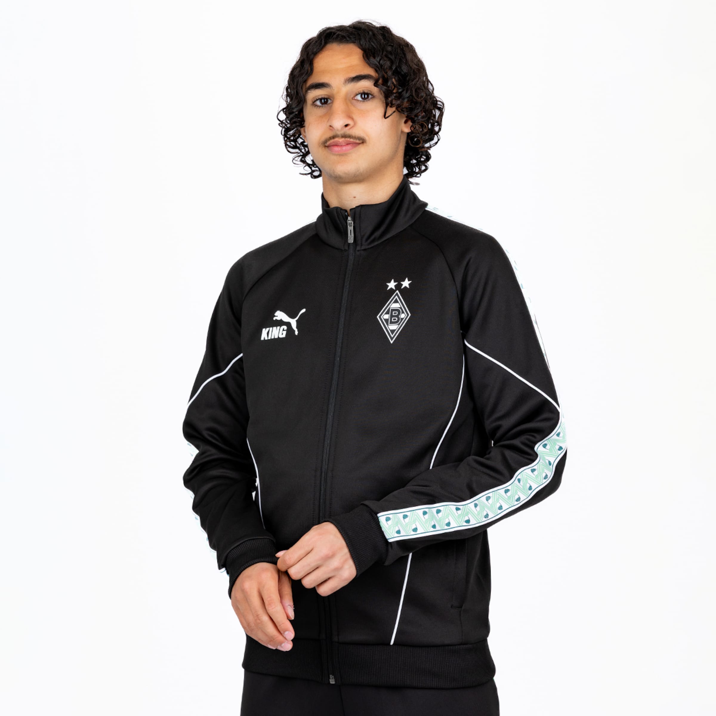 Junger Mann mit lockigem Haar in schwarzer Puma Borussia Mönchengladbach Trainingsjacke vor weißem Hintergrund.