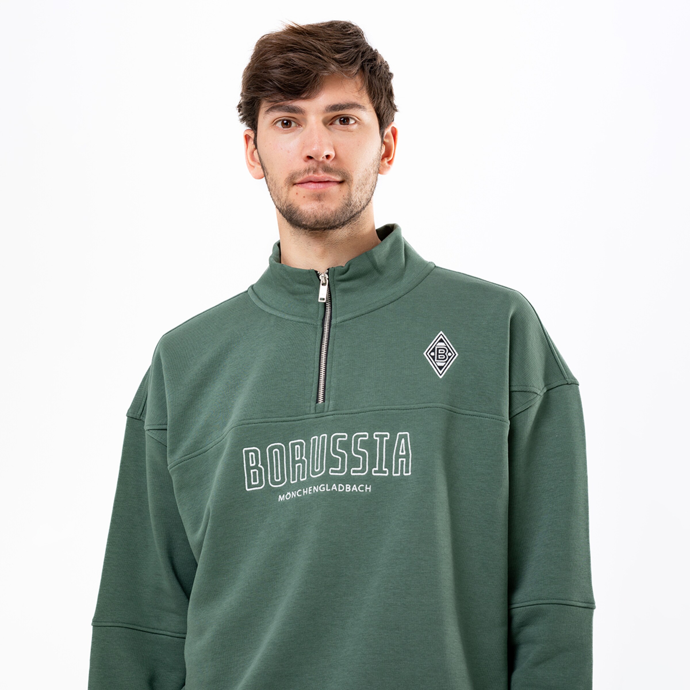 Mann in grünem Borussia Mönchengladbach Sweatshirt mit Stehkragen, Reißverschluss, Vereinslogo und weißem Schriftzug vor weißem Hintergrund.