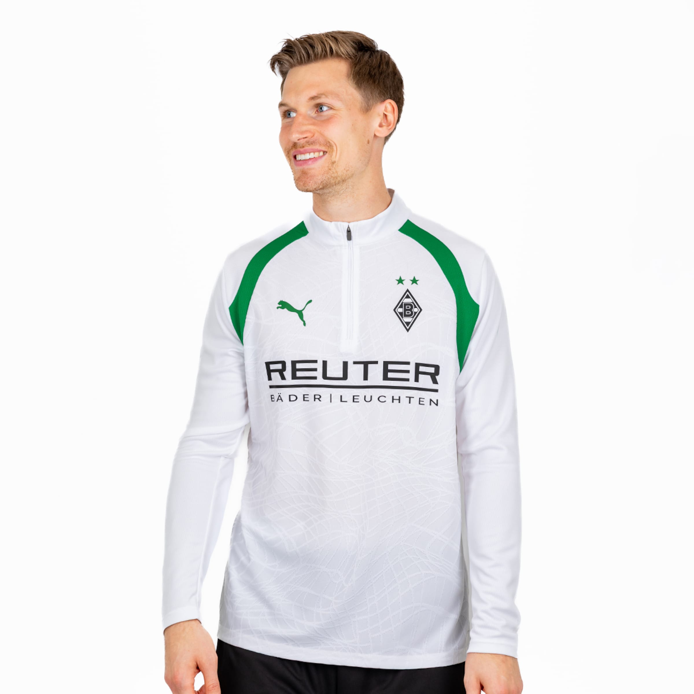 Mann im weißen Langarm-Trainingsshirt von Borussia Mönchengladbach mit grünen Akzenten. Es zeigt das Puma-Logo, das Vereinswappen mit zwei Sternen und den Sponsor REUTER. Er lächelt in die Seite.