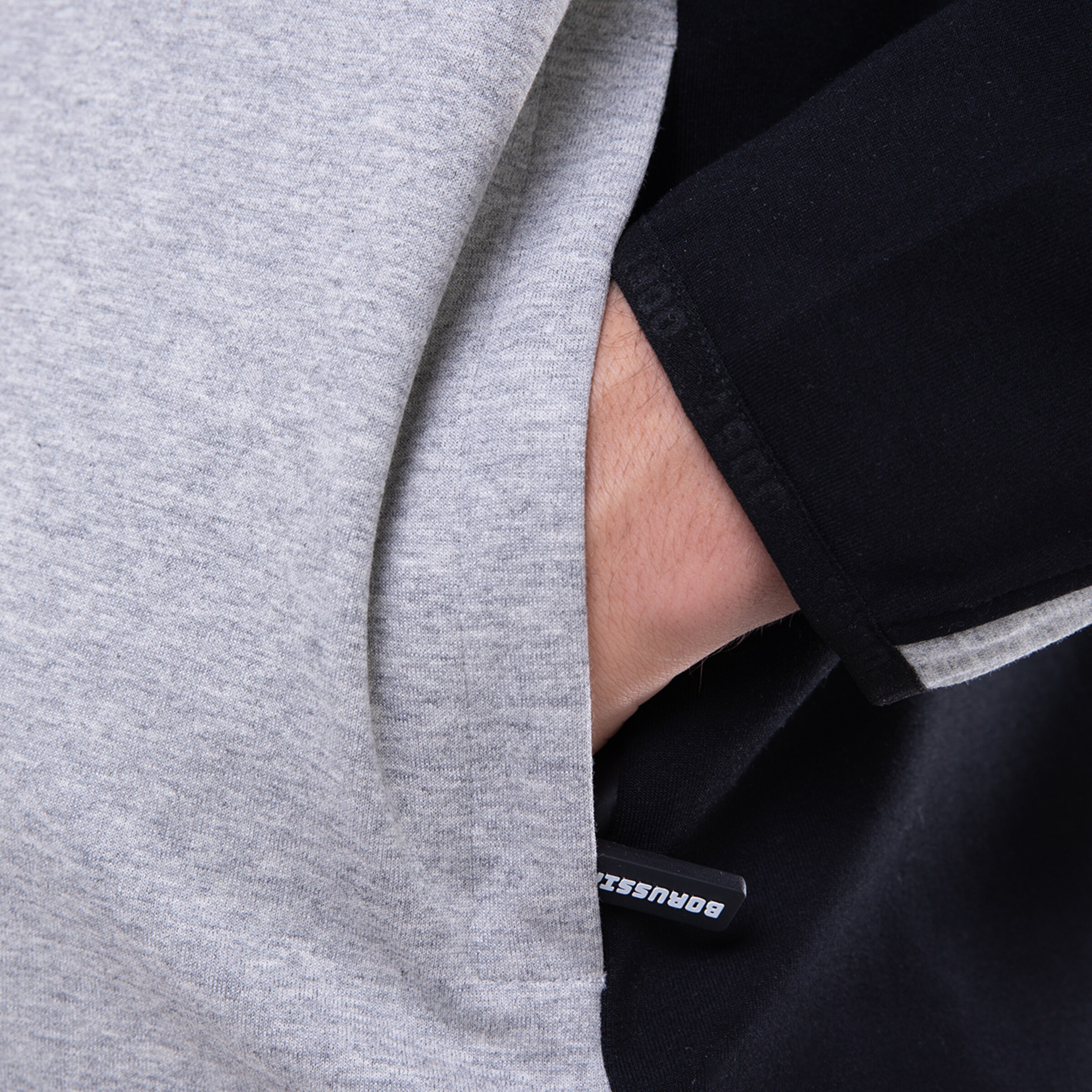 Detailansicht eines grauen Shirts mit schwarzem Ärmel und aufgesticktem Logo.