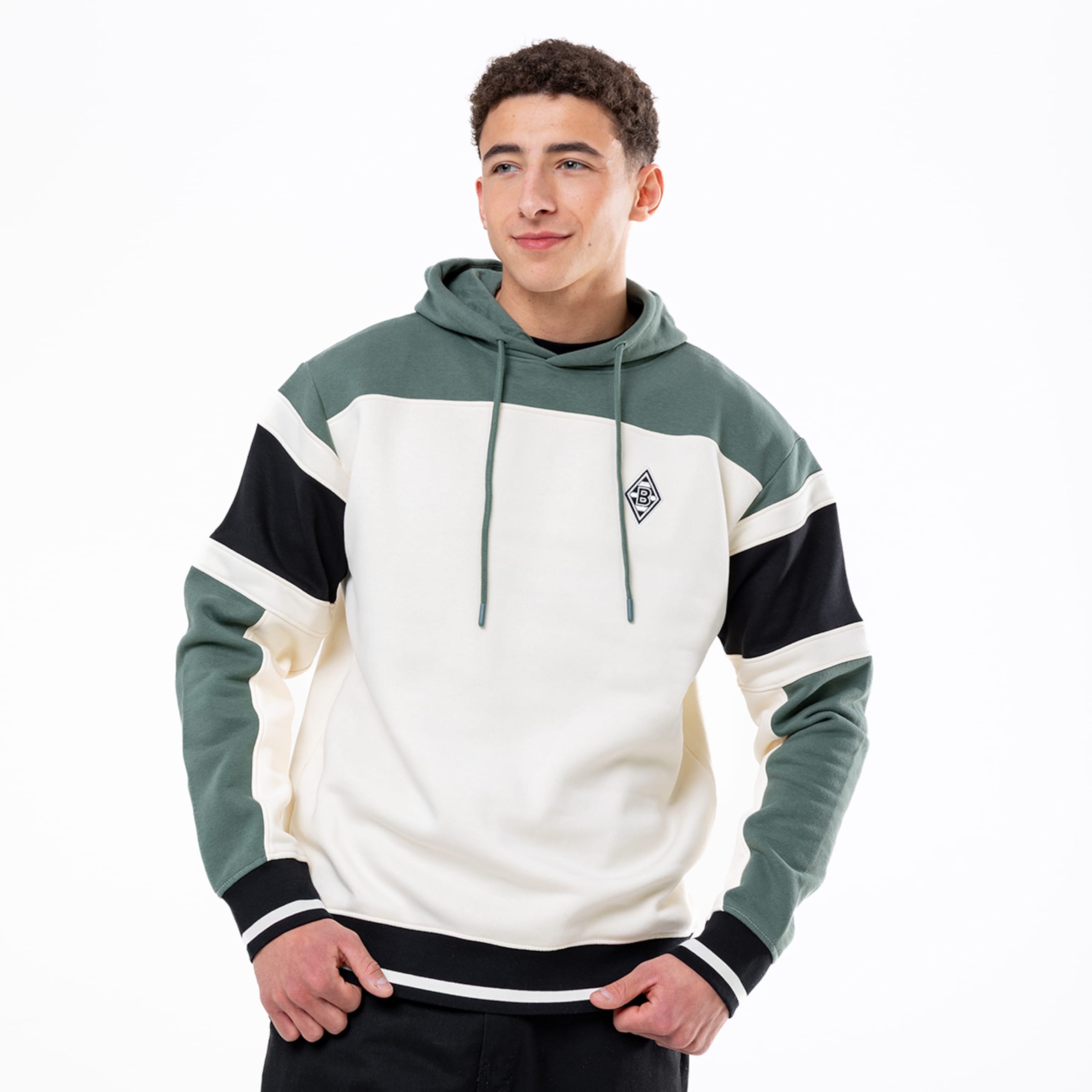 Junger Mann in einem grün-weiß-schwarzen Hoodie mit Colourblocking und kleinem Logo vor einem weißen Hintergrund.