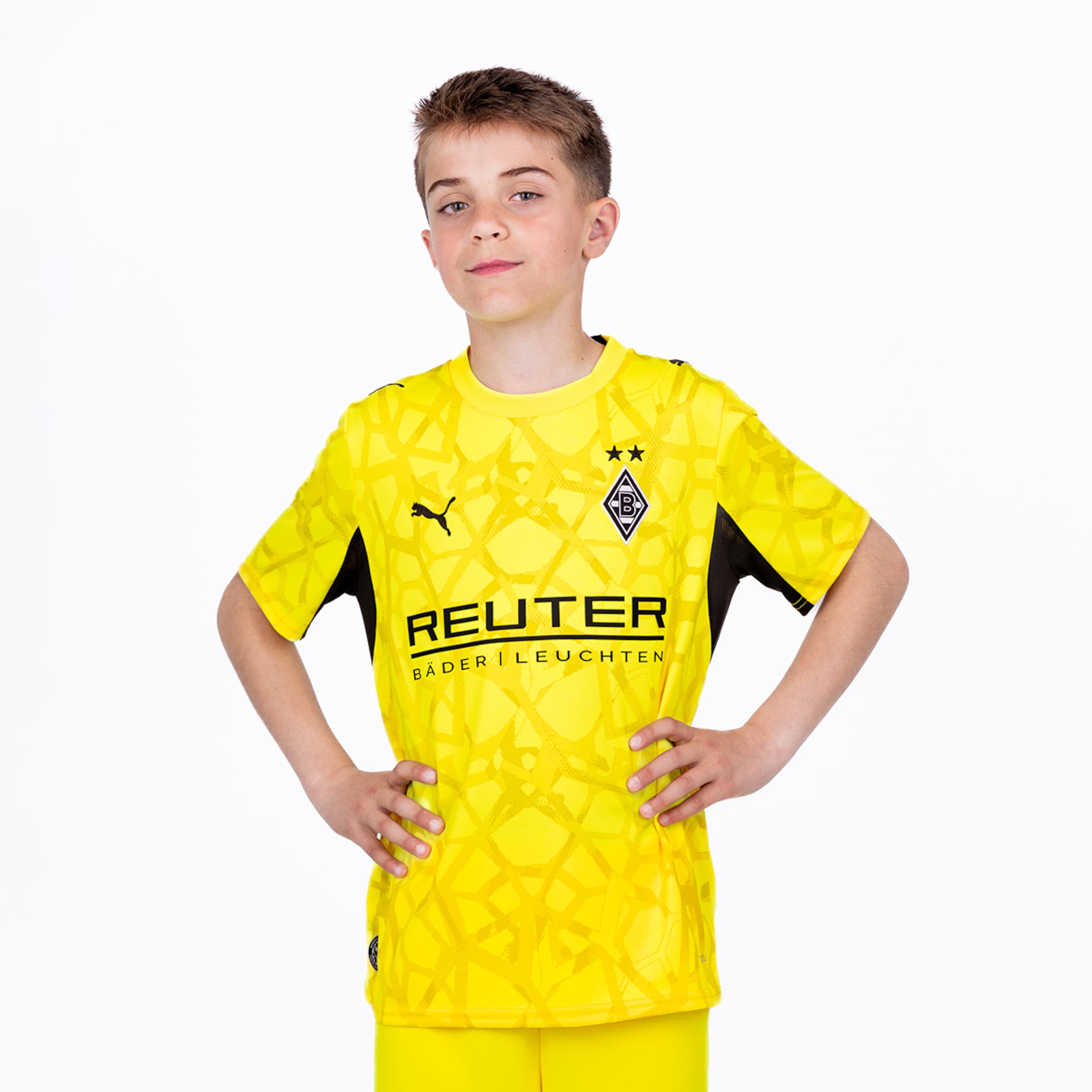 Ein Junge im gelben Gladbach-Torwarttrikot mit Reuter-Sponsor und Puma-Logo, Hände an den Hüften vor weißem Hintergrund.