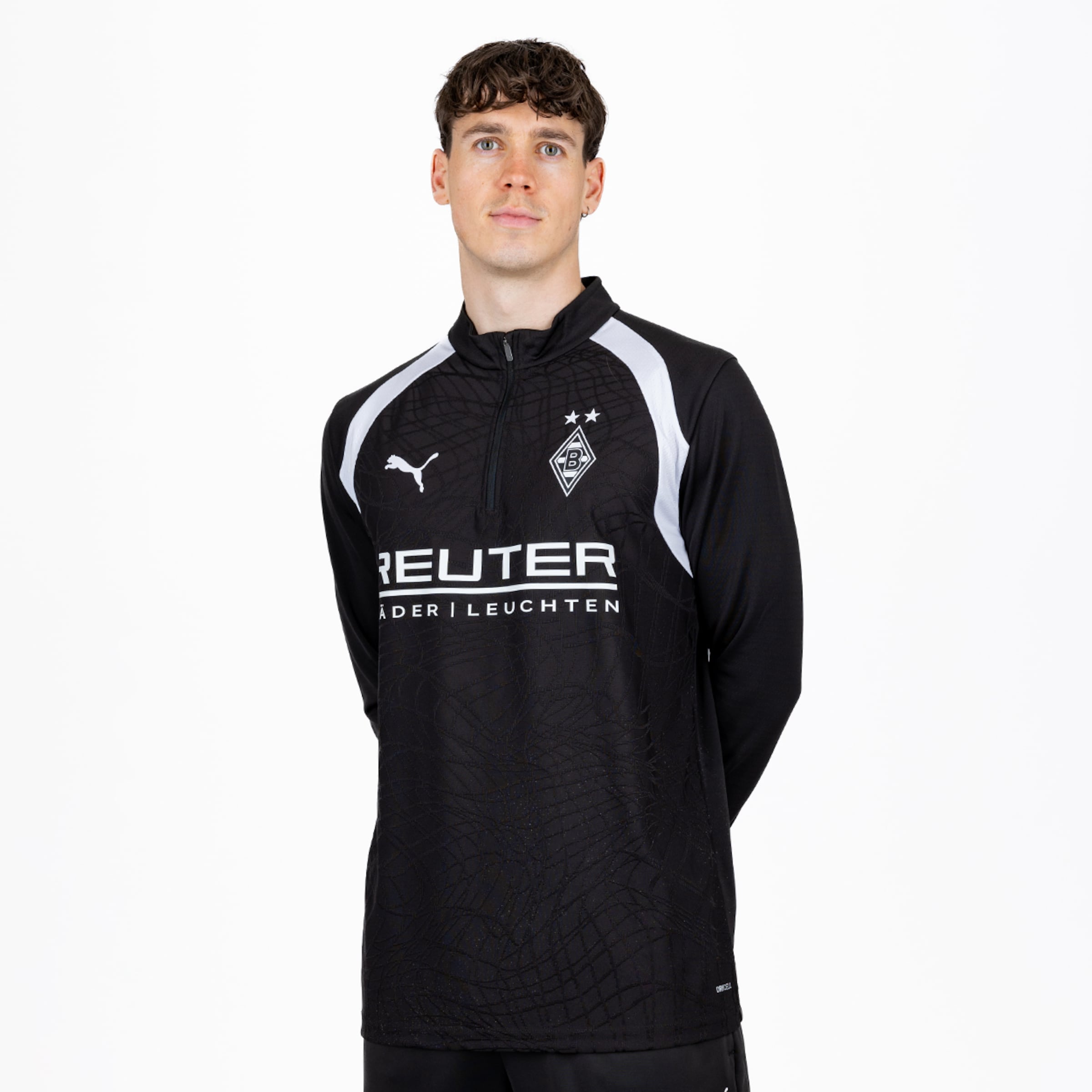 Ein junger Mann mit lockigem braunem Haar und hellen Augen steht vor weißem Hintergrund. Er trägt ein schwarzes Borussia Mönchengladbach Trainingsshirt mit weißen Akzenten, Puma-Logo, Vereinswappen und „REUTER“-Sponsor.