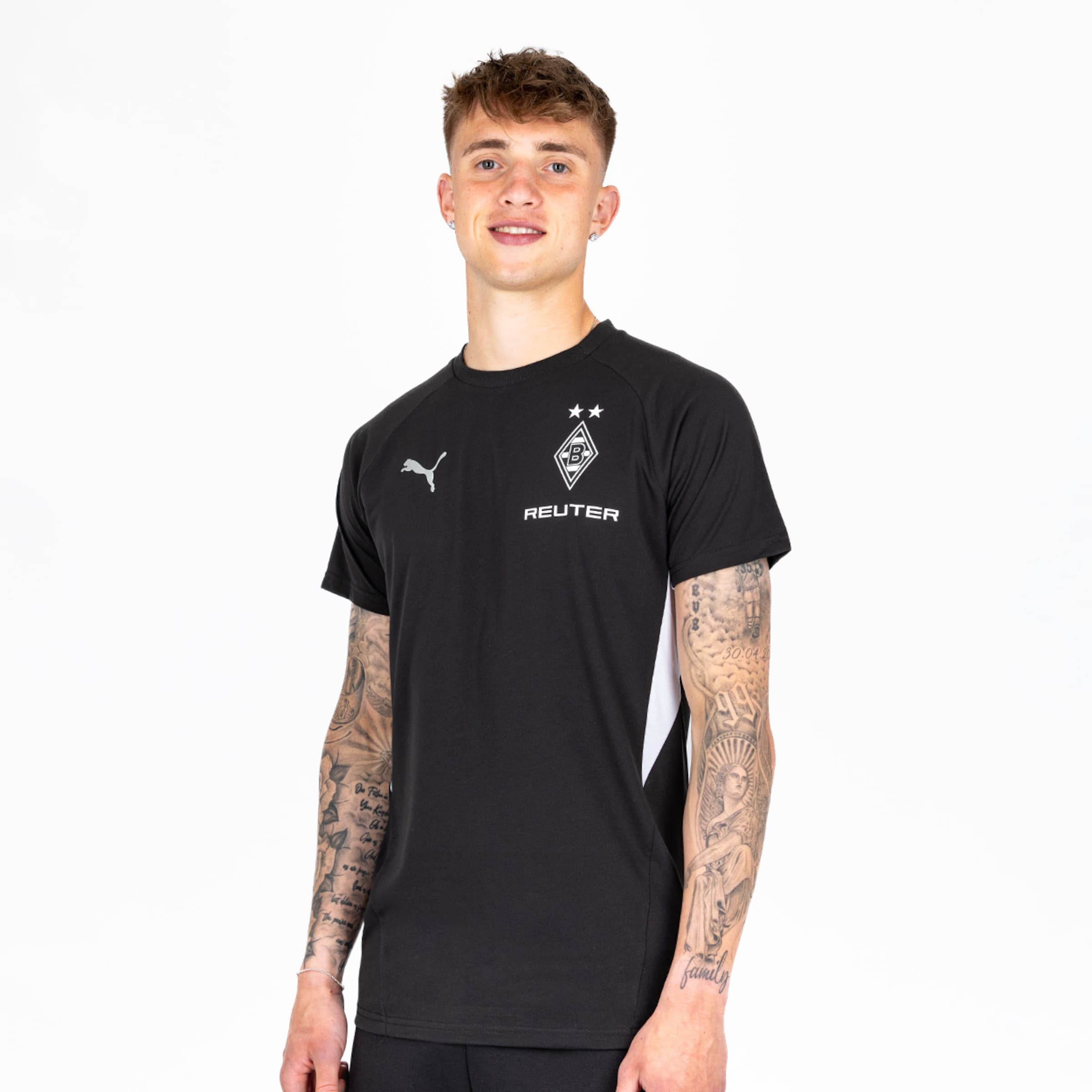 Ein junger Mann mit tätowierten Armen trägt ein schwarzes Fußballtrikot mit dem Logo von Borussia Mönchengladbach.