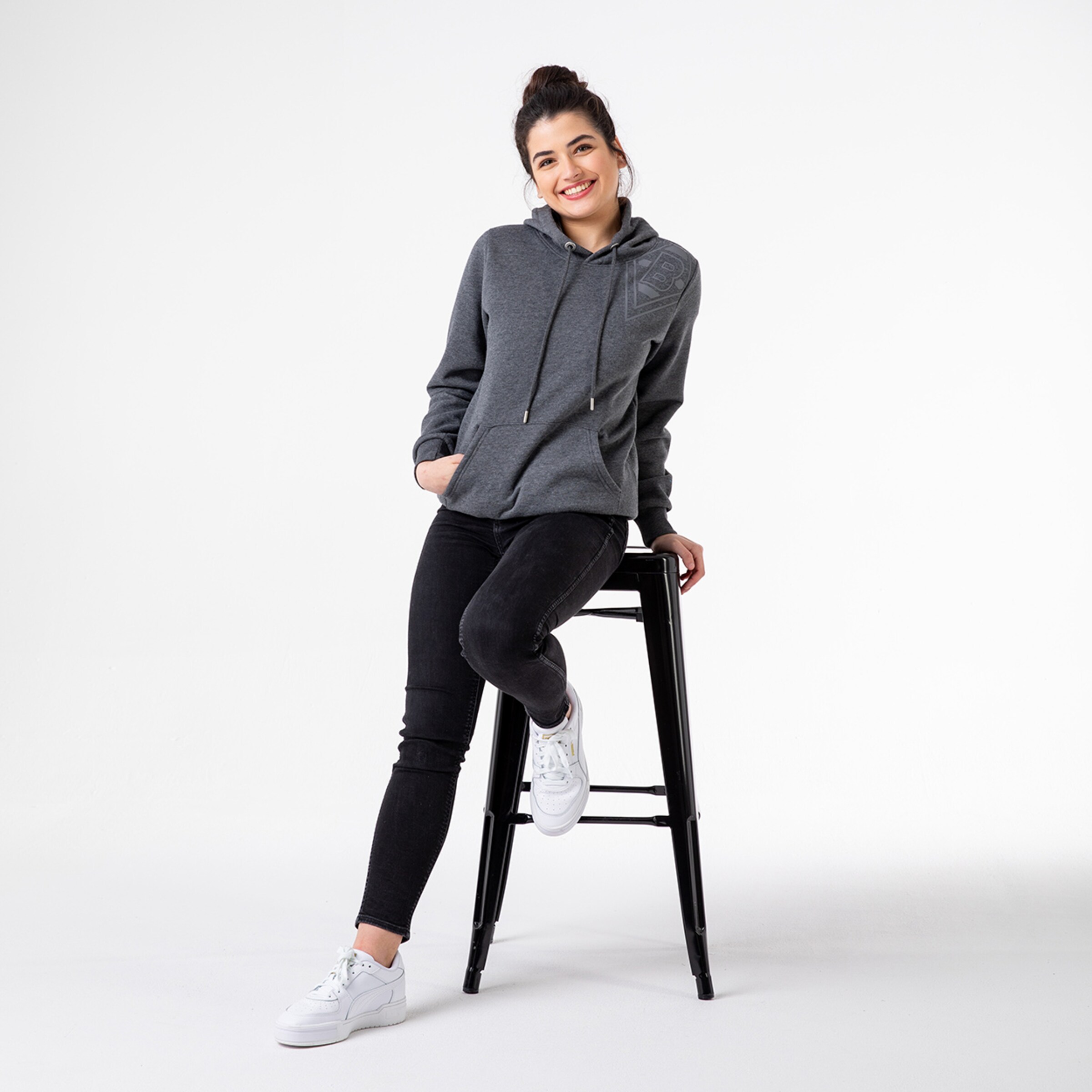 Junge Frau in grauer Kapuzenjacke sitzt lächelnd auf einem Hocker.