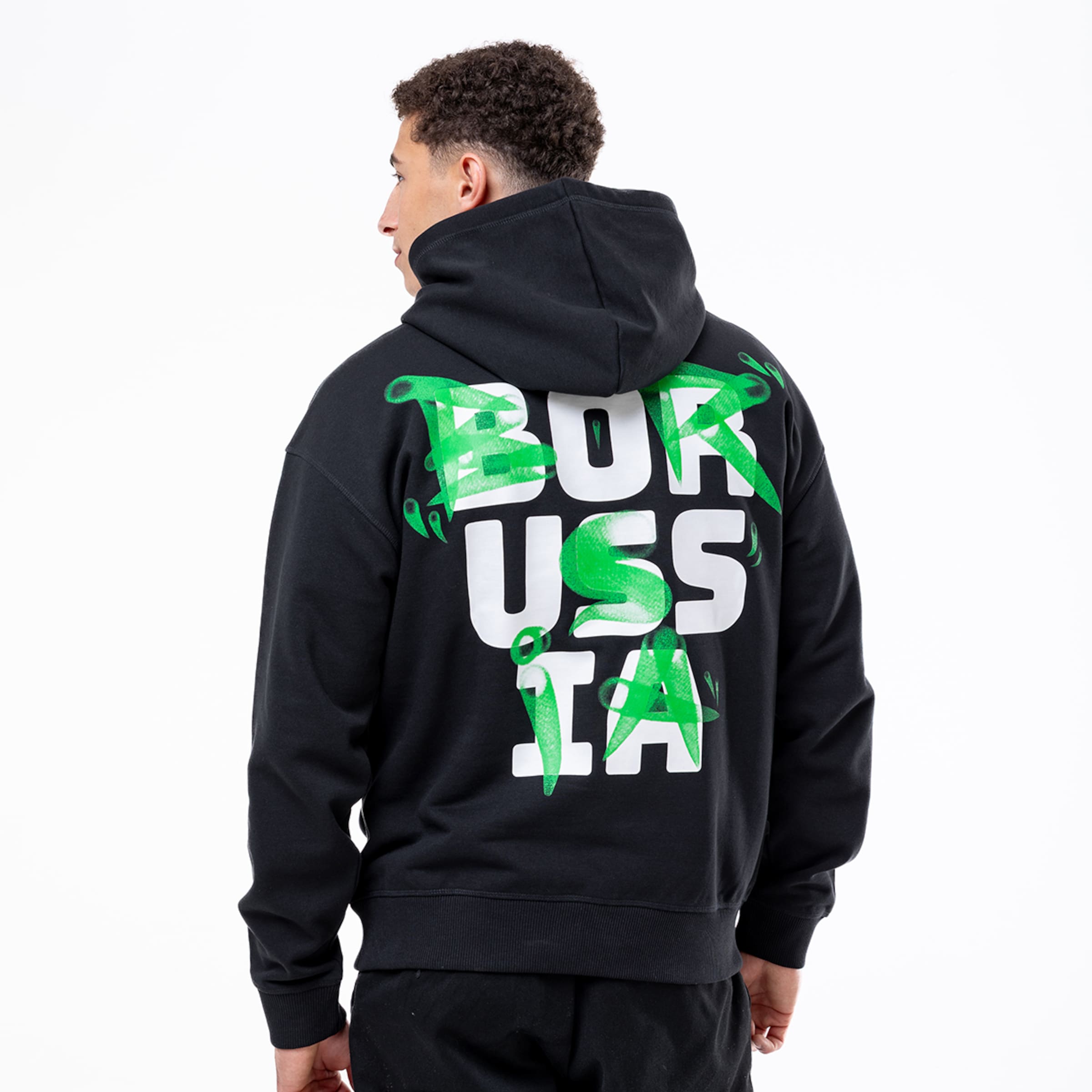 Rückansicht eines Mannes im schwarzen Hoodie mit weißem „BORUSSIA“-Schriftzug und grünen Graffiti-Akzenten vor weißem Hintergrund.