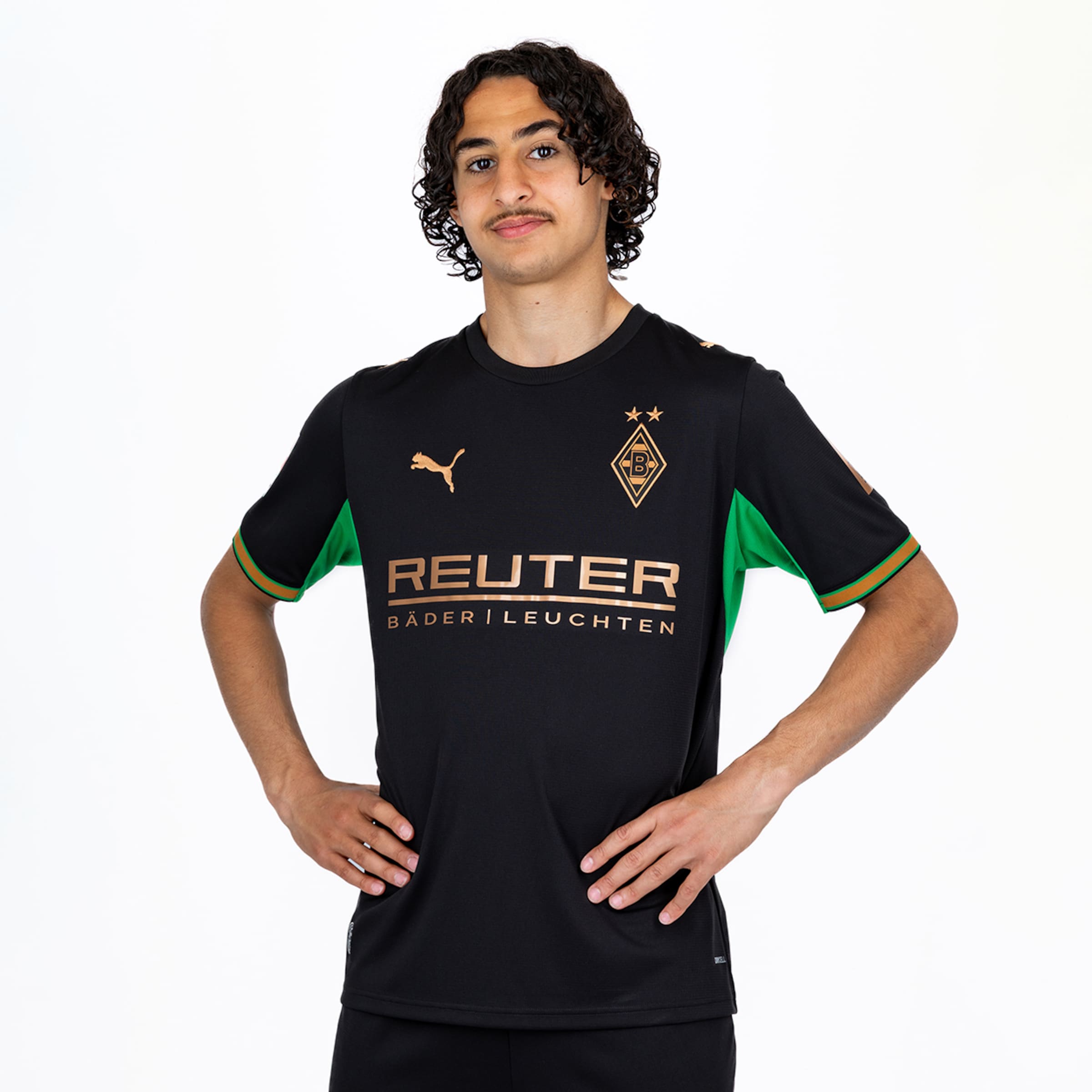 Junger Mann mit dunklen Locken und Schnurrbart im schwarzen Borussia Mönchengladbach Trikot, Hände in die Hüften gestemmt, vor weißem Hintergrund.