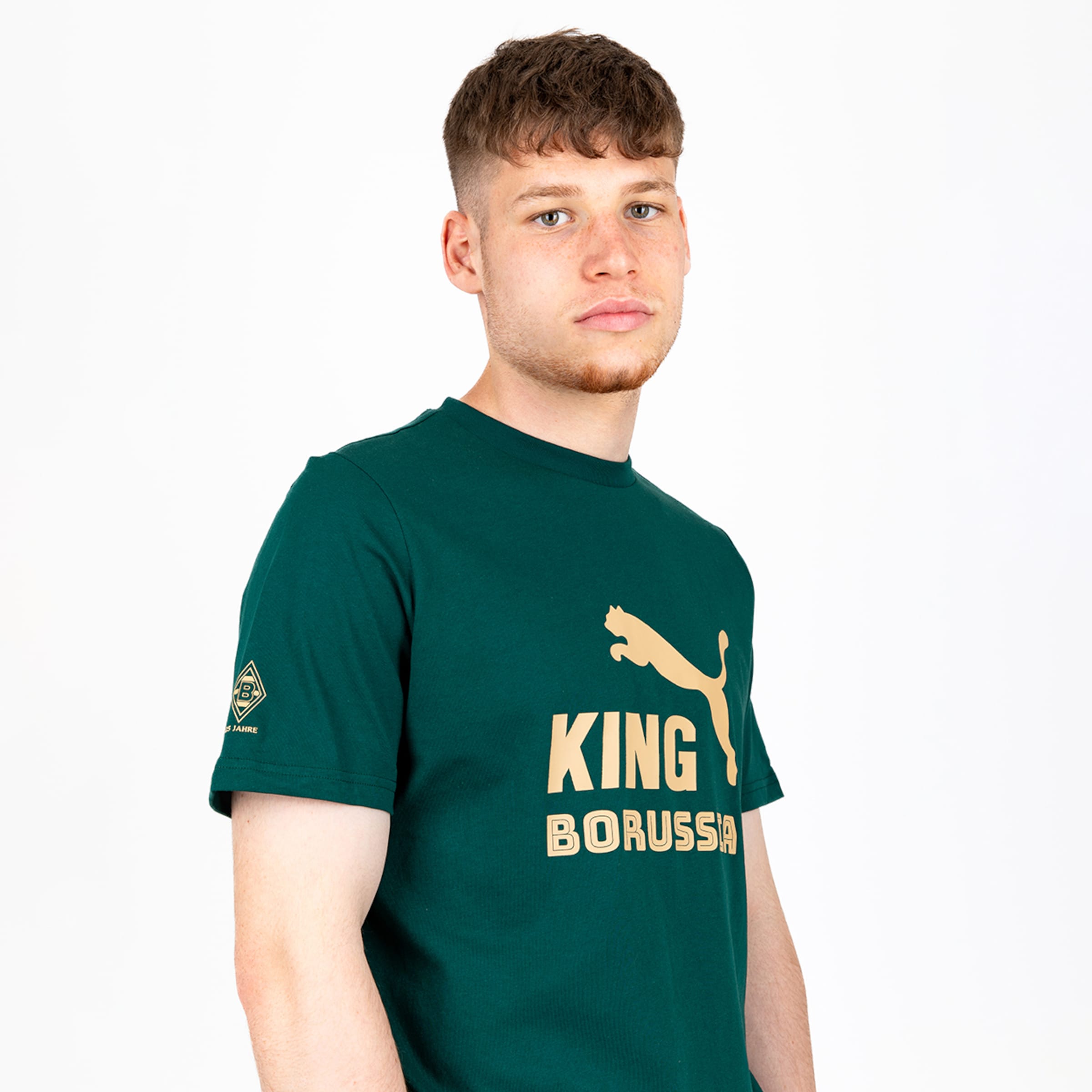 Junger Mann in dunkelgrünem Puma T-Shirt mit goldenem „KING BORUSSIA“ Print und Vereinslogo am Ärmel vor weißem Hintergrund.