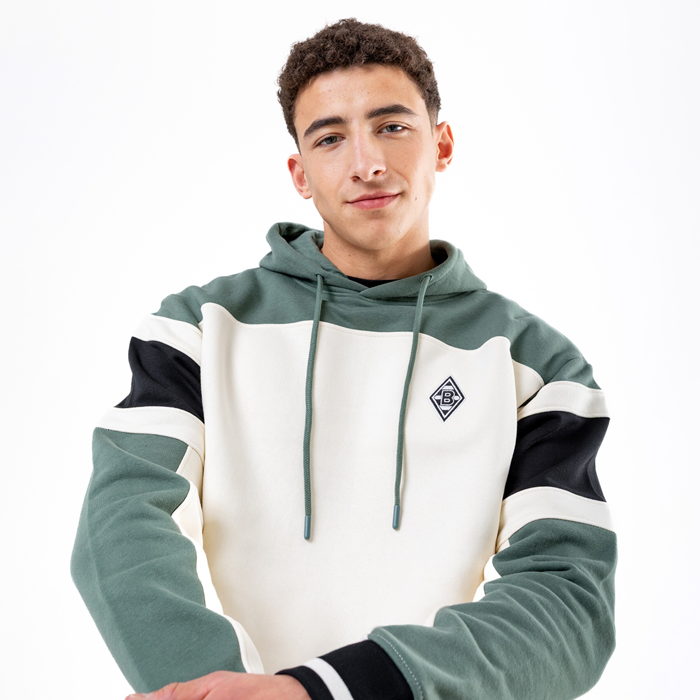 Junger Mann mit lockigem Haar trägt einen grün-weiß-schwarzen Hoodie mit Rautenlogo vor weißem Hintergrund.