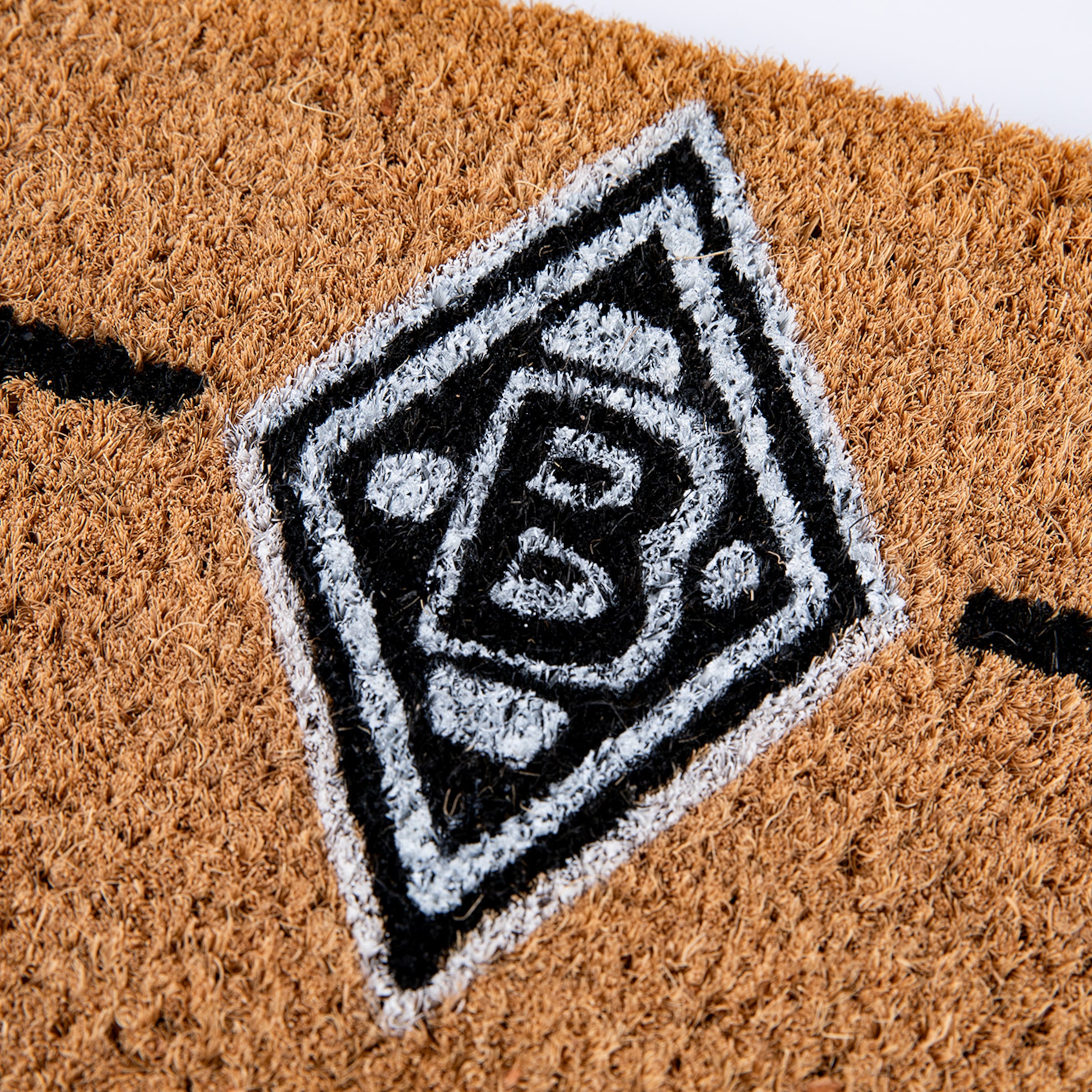 Nahaufnahme einer Fußmatte aus Kokosfasern mit dem schwarz-weiß-gemalten Logo von Borussia Mönchengladbach in Rautenform, umgeben von braunen Fasern.