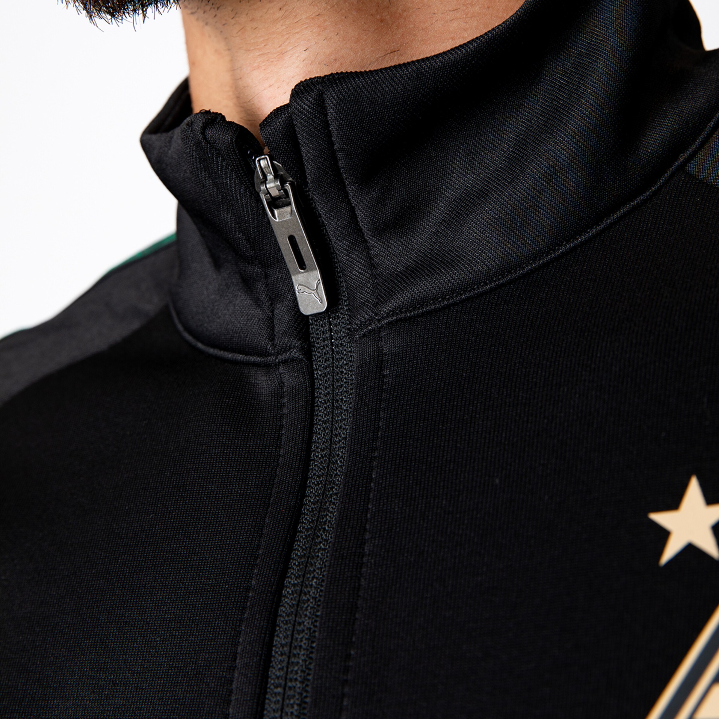 Nahaufnahme einer schwarzen Puma Trainingsjacke mit Stehkragen und Metall-Reißverschluss mit Logo. Ein goldener Stern ist auf der Brust erkennbar.