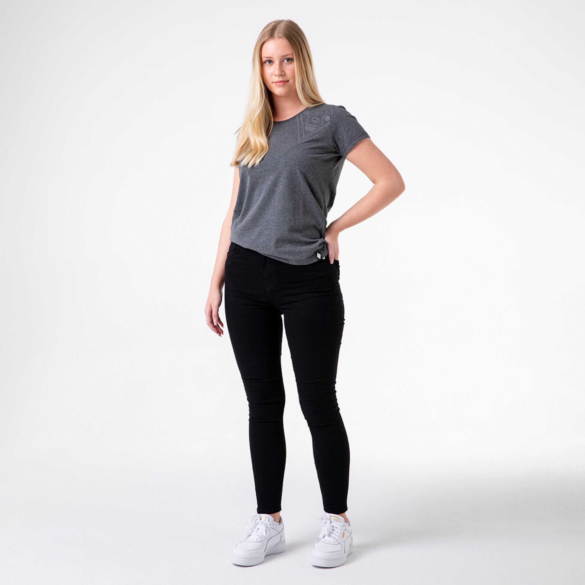 A woman with blonde hair wears a gray t-shirt and black skinny jeans standing with one hand on her hip.