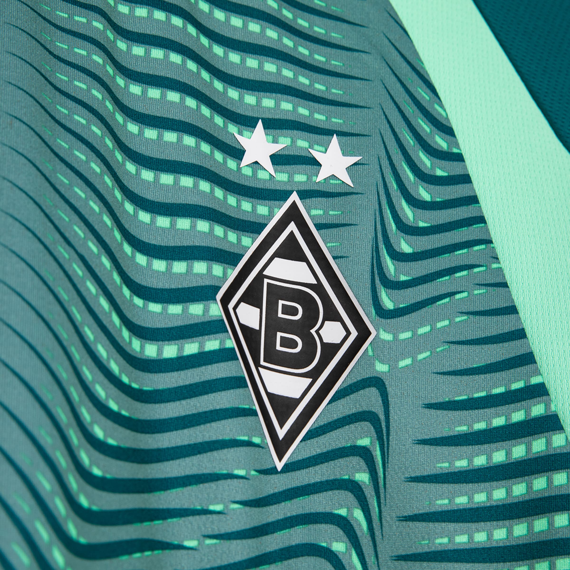 Close-up of a mint green jersey with a wave pattern, dark green stripes, and light green rectangles. The Borussia Mönchengladbach crest and two white stars are visible. A section of a mint green mesh side panel is on the right.