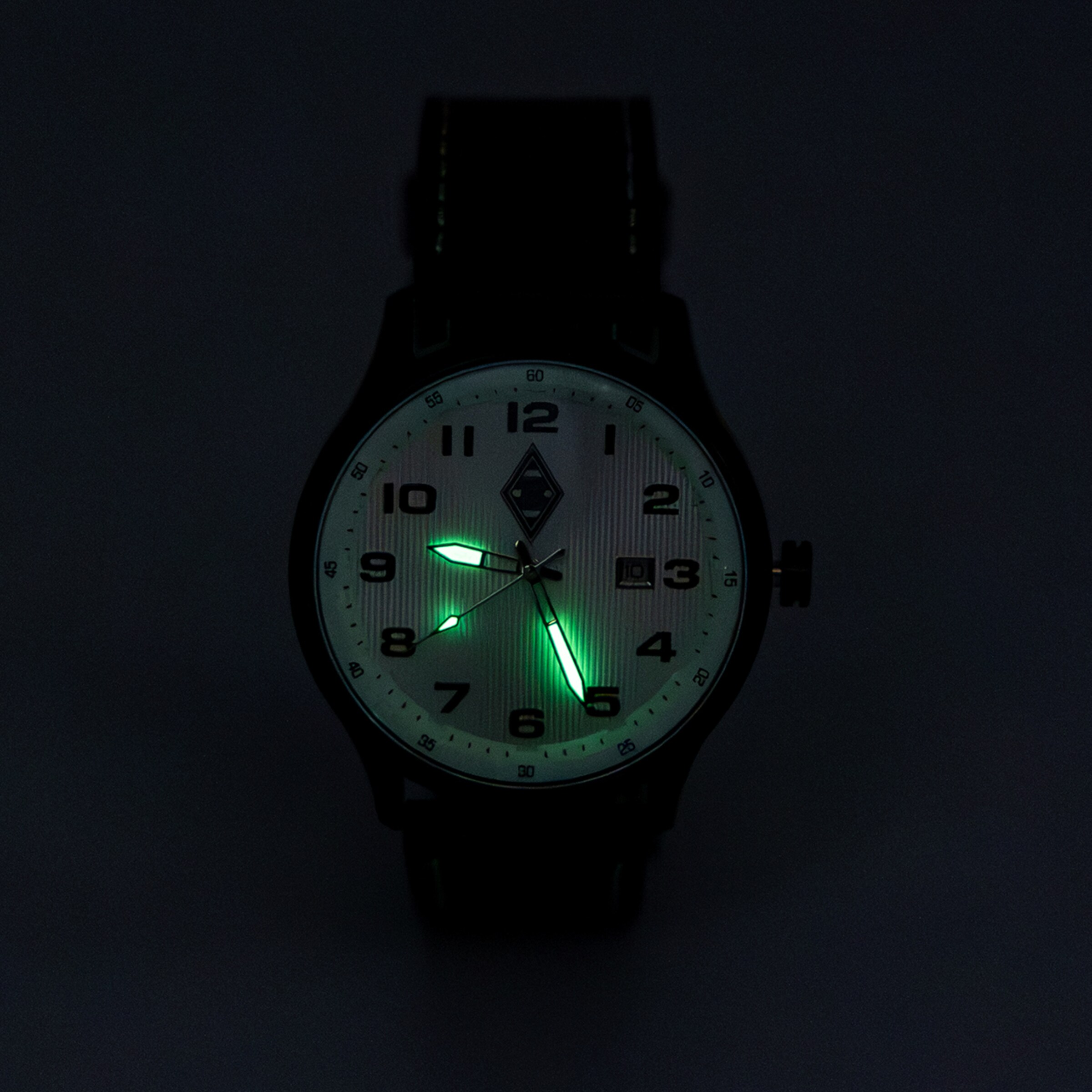 Armbanduhr im Dunkeln mit grün leuchtenden Zeigern auf hellem Zifferblatt mit schwarzen Ziffern. Schwarzes Gehäuse und Armband.