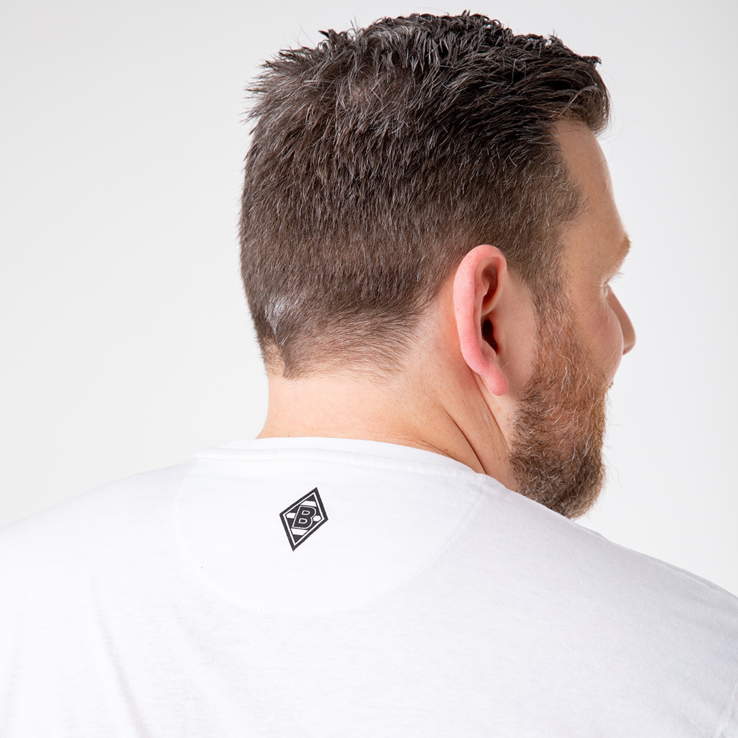 Mann mit kurzem grauem Haar von hinten trägt ein weißes T-Shirt mit einem kleinen Logo auf dem Nacken.